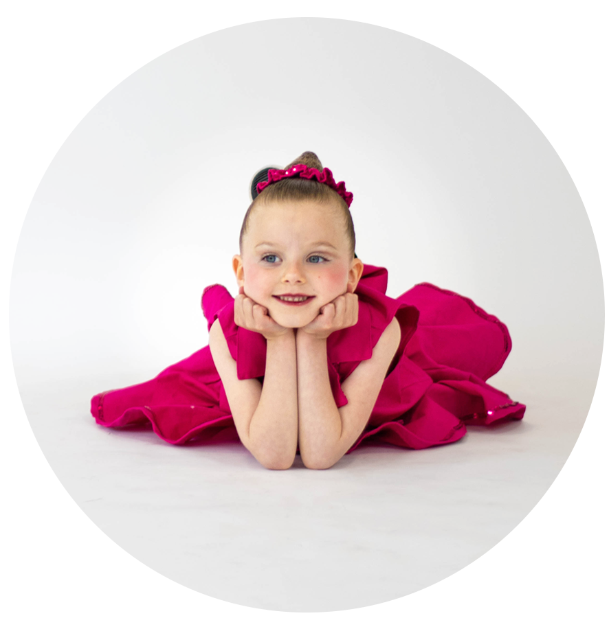 Young girl in a pink dress laying on her stomach with chin resting on her hands, smiling, with a plain white background.