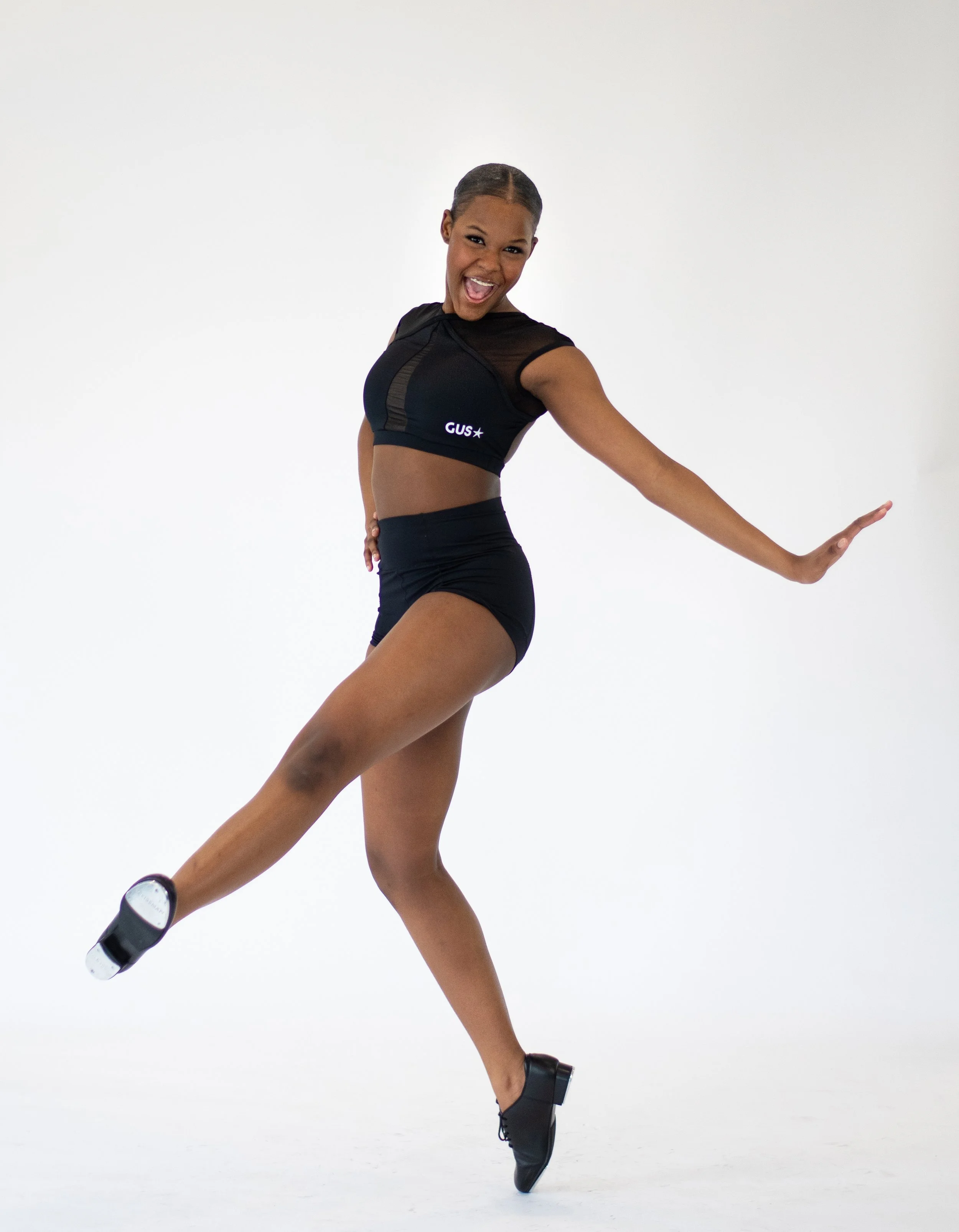 A young tap dancer smiling and posing against a white background, wearing black athletic clothing.