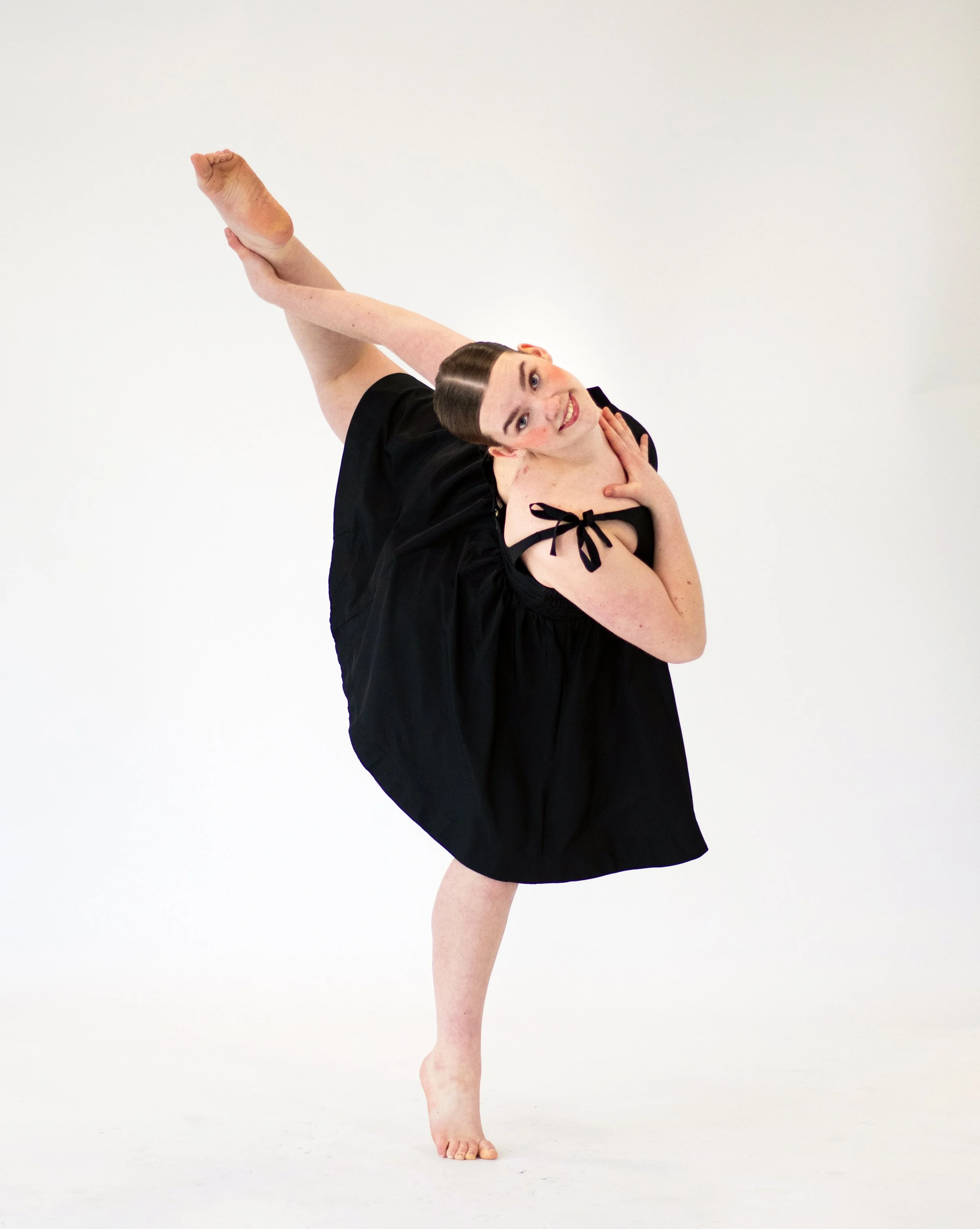 A dancer with one leg raised and bent backward, holding the foot with her hand, wearing a black dress, in a dance studio.