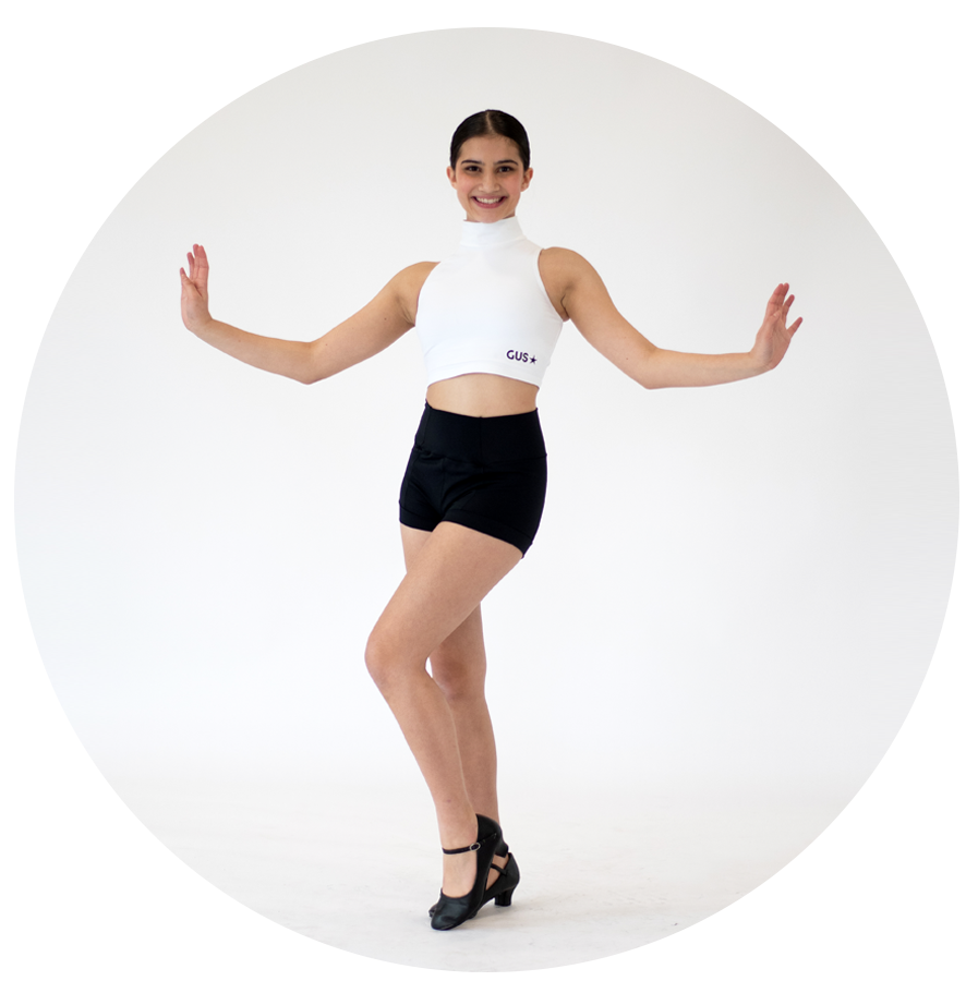 A young woman in black dance shoes, a white sleeveless crop top, and black shorts poses against a plain white background with a big smile.