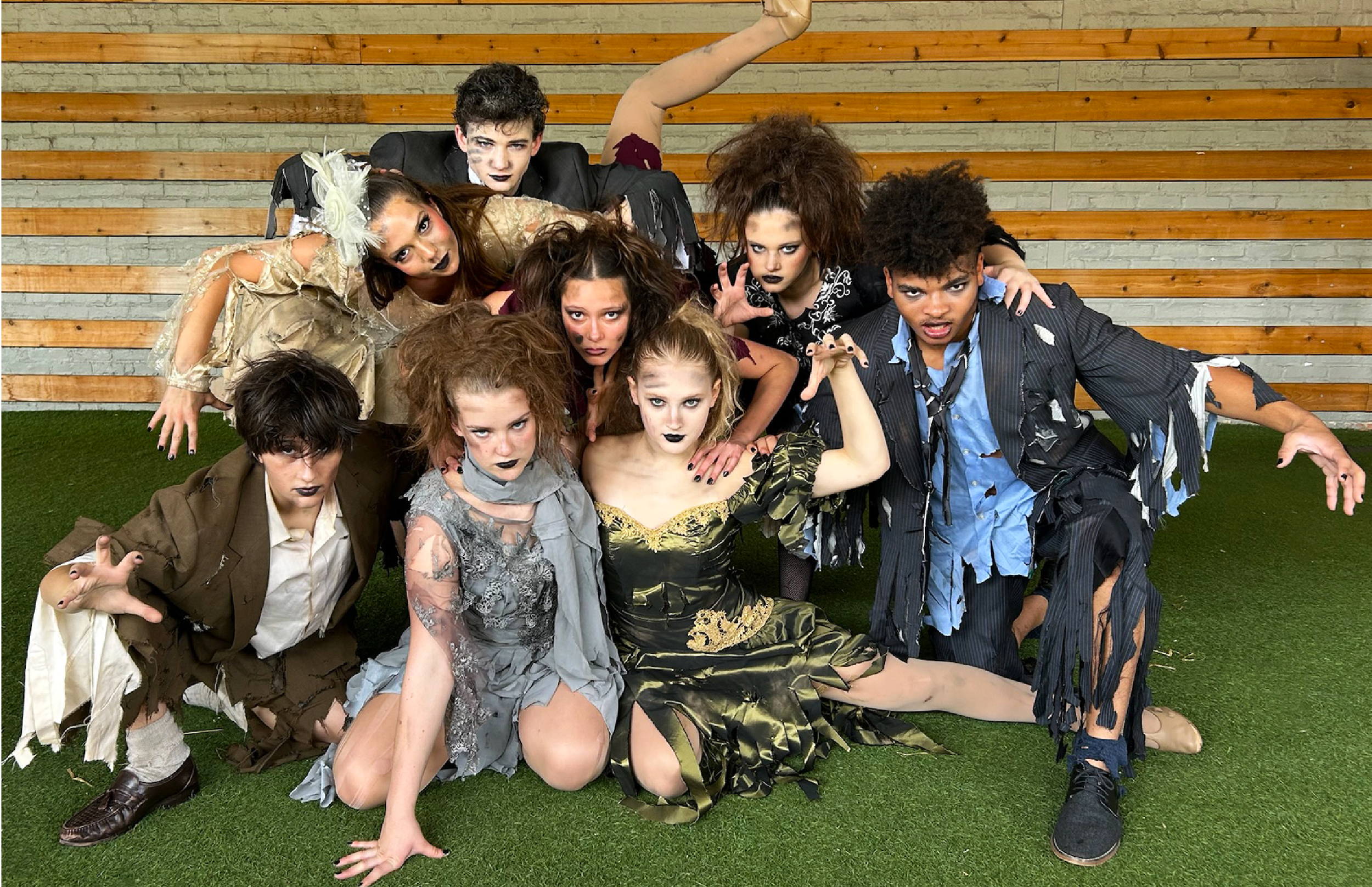 A group of ten individuals dressed in tattered, zombie-like costumes, posed in a theatrical or Halloween-themed scene in front of a brick and wood wall. They have intense facial expressions, dark makeup around their eyes and lips, and are crouched or leaning forward as if stalking or attacking.