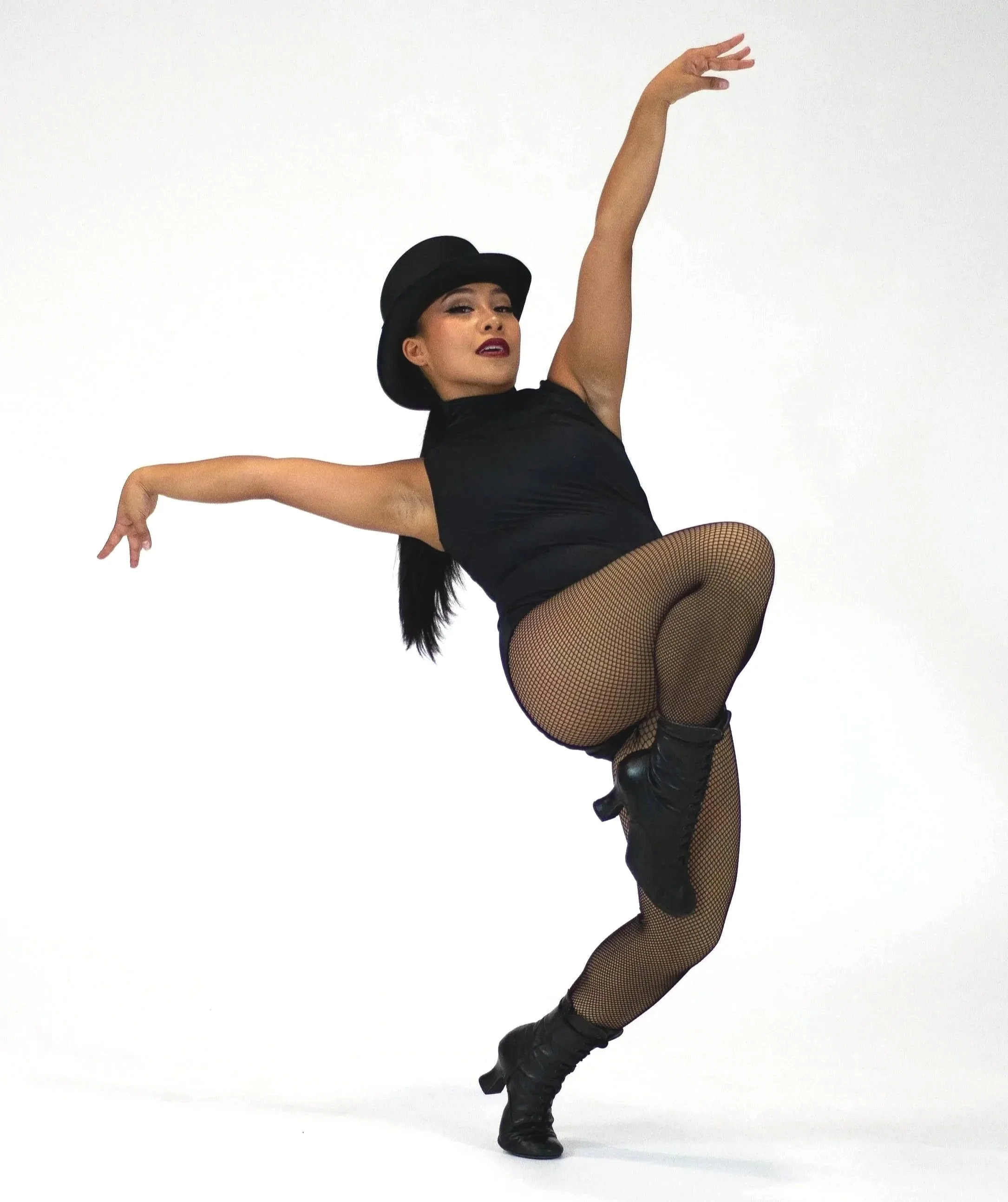 A woman performing a dance pose wearing a black hat, black top, fishnet stockings, and black high-heeled boots against a plain white background.