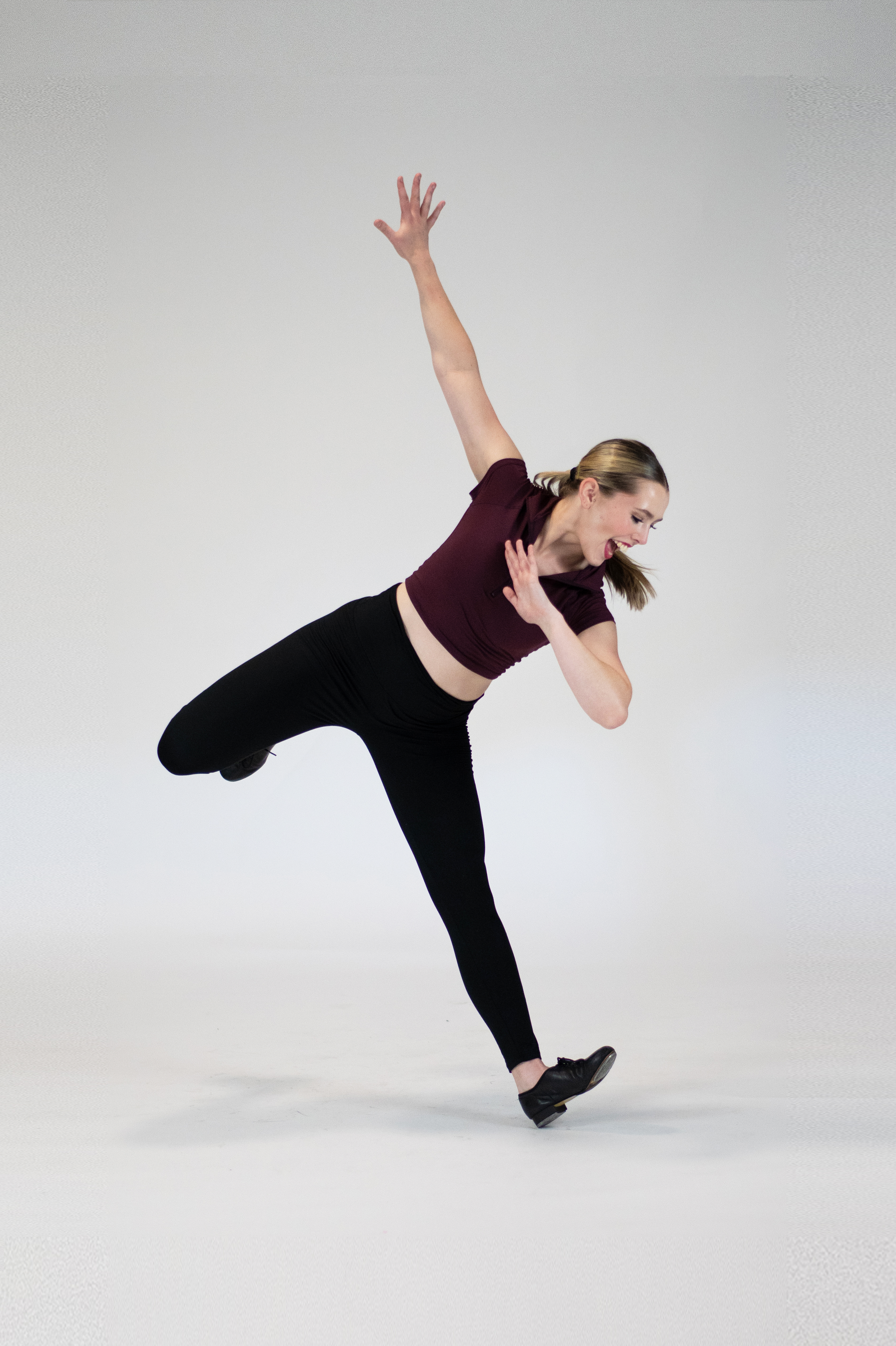 A tap dancer with one arm raised above her head and the other near her chest. She is wearing a maroon crop top, black leggings, and black shoes, with her hair pulled back.