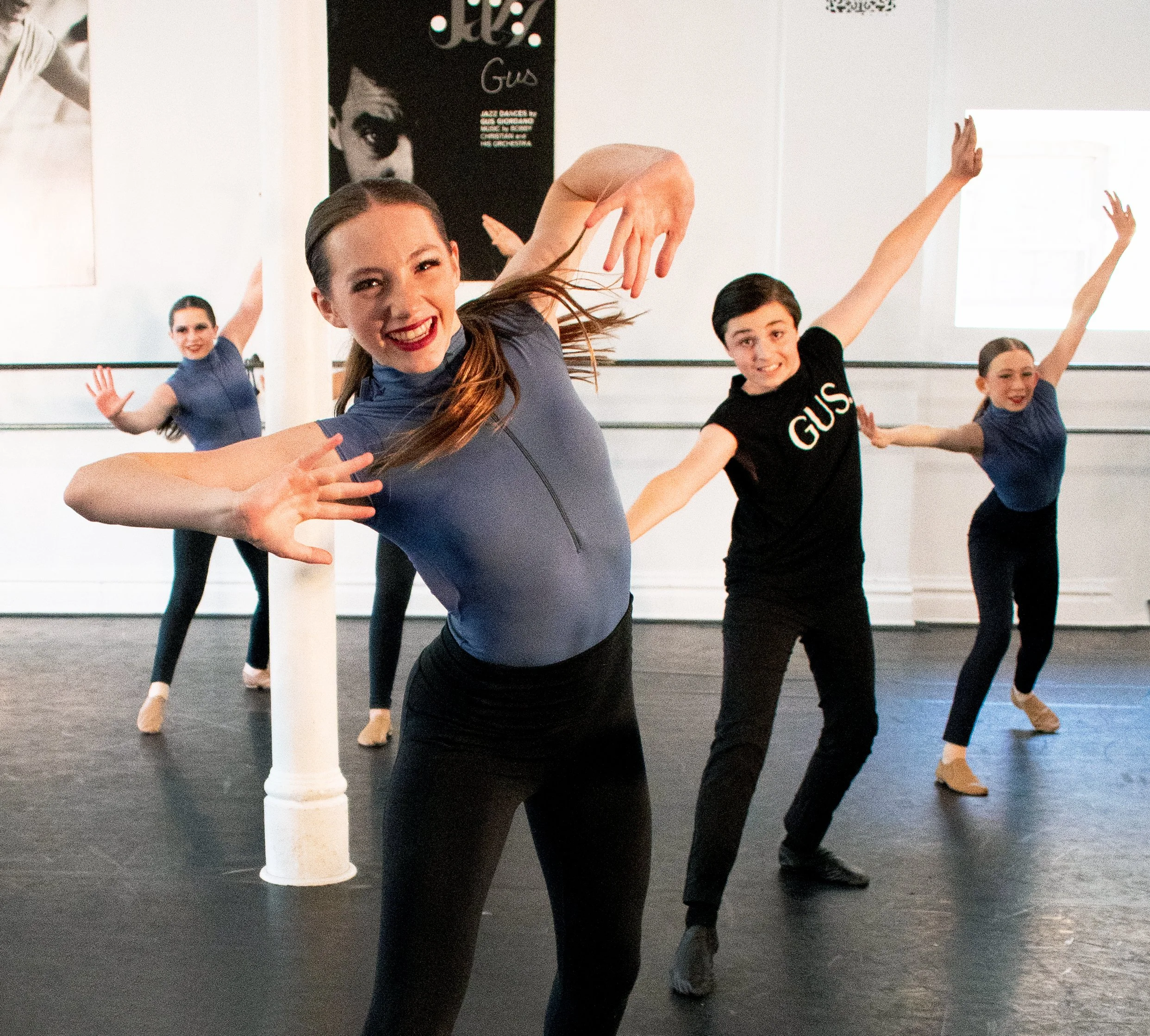 A group of five ballet dancers practicing in a dance studio, with the instructor in the foreground smiling and stretching.