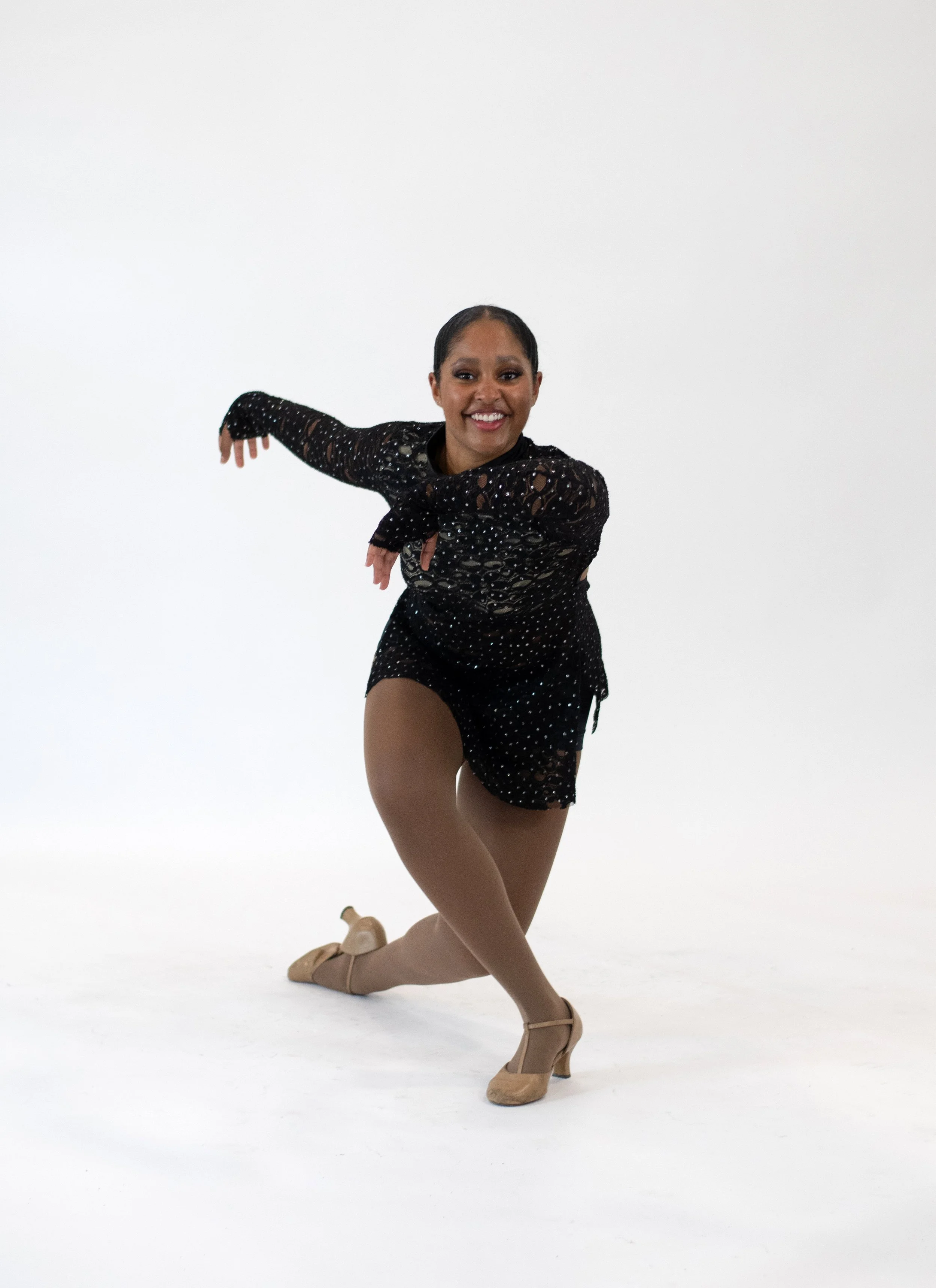 A female jazz dancer posing on a white background, wearing a black lace costume with sequins, tan tights, and character shoes.