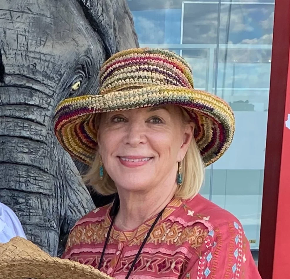 A woman smiling, wearing a multicolored straw hat and a vibrant, red patterned top, standing next to an elephant statue in a zoo or similar setting.