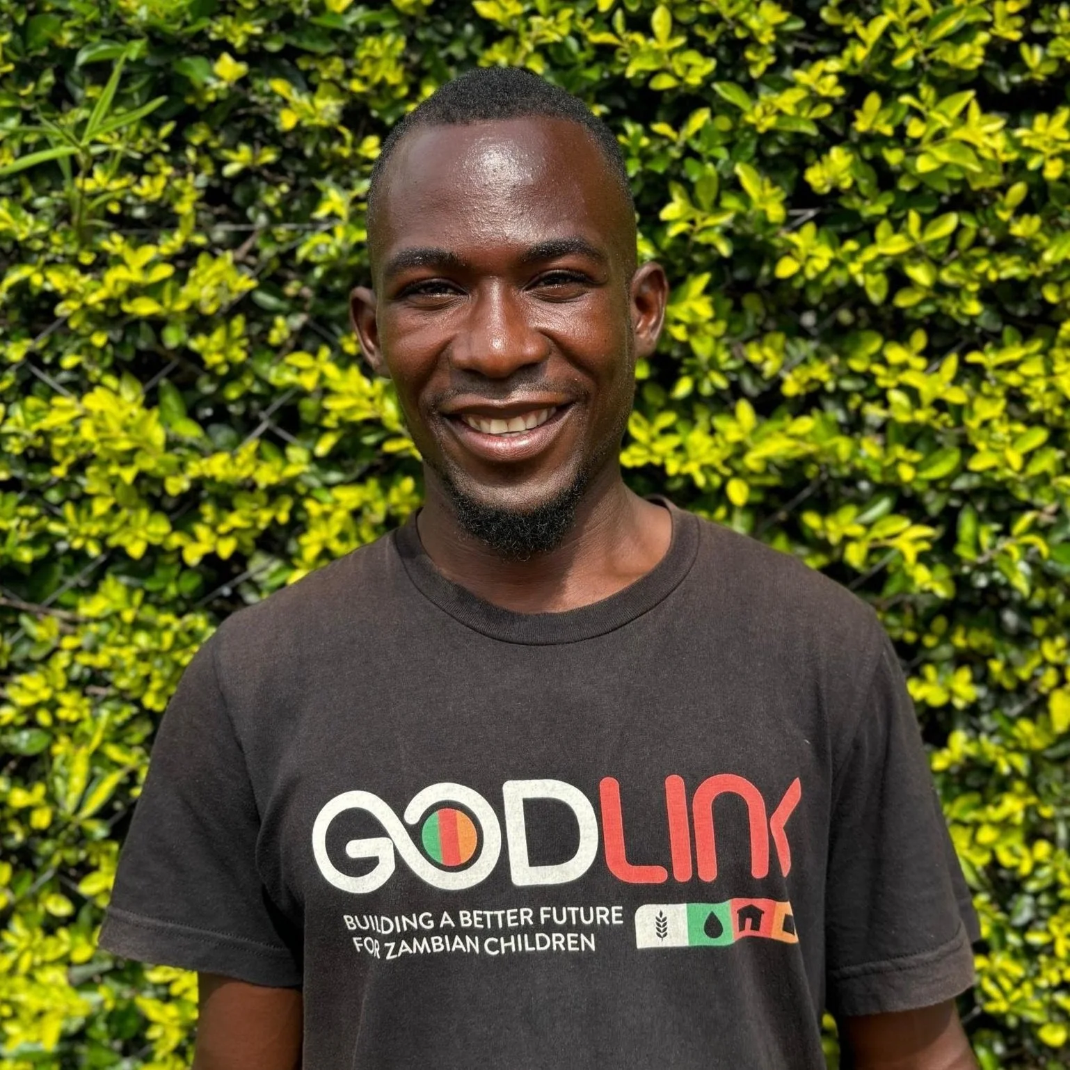 A smiling man with short hair and a beard stands in front of green foliage, wearing a black T-shirt with the logo 'GODLINK' and the slogan 'Building a better future for Zambian children'.