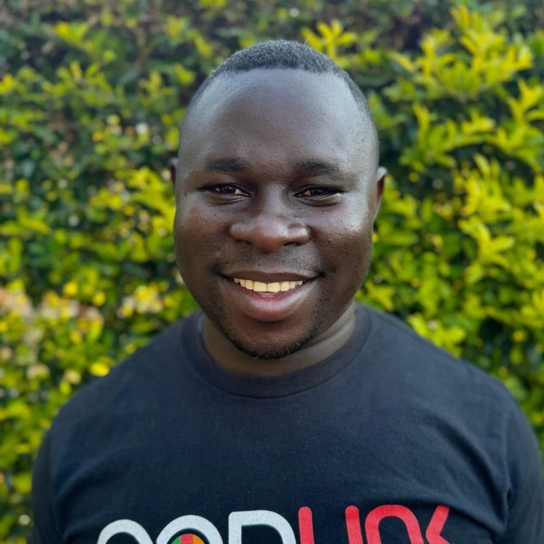 A smiling man with short hair and a goatee, wearing a black t-shirt with partially visible colorful text, standing outdoors with green foliage in the background.