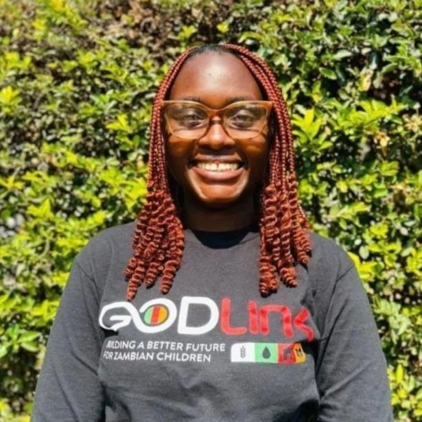 A woman with braids and glasses standing outdoors in front of green foliage, smiling, wearing a dark T-shirt with the logo 'GODLink' and text supporting Zambian children.