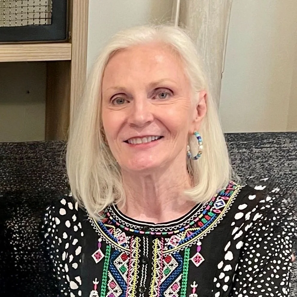 A smiling elderly woman with long white hair, wearing a black embroidered blouse with colorful patterns and hoop earrings, sitting on a patterned couch in a cozy indoor setting.