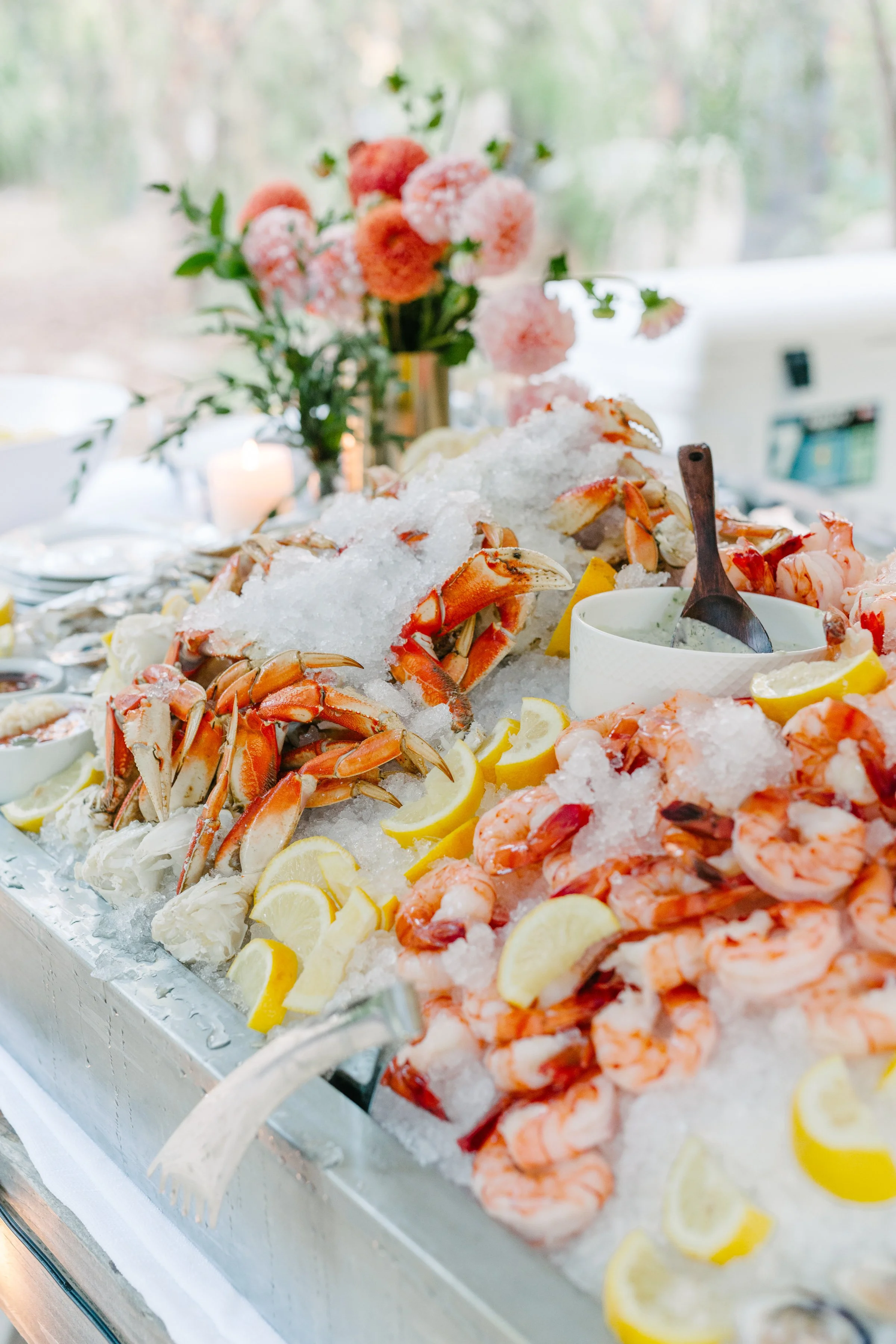 Seafood display on ice with lobster, shrimp, lemon slices, and sauce, with pink flowers in the background.