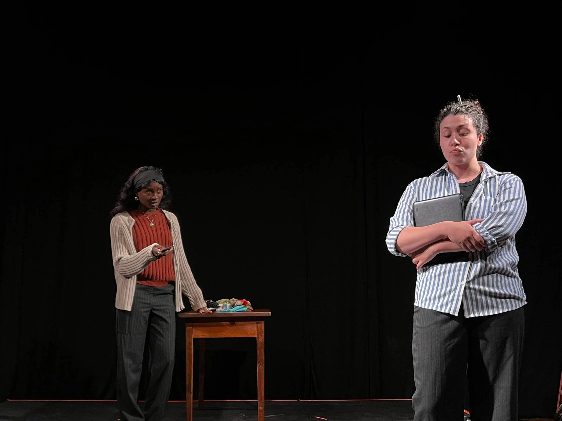 Two women having a conversation on stage, with dark curtains in the background. The woman on the left has blonde hair and a distressed expression, wearing a maroon top. The woman on the right has dark hair in a ponytail, wearing earrings, a black blazer, and a red lace top, speaking animatedly.