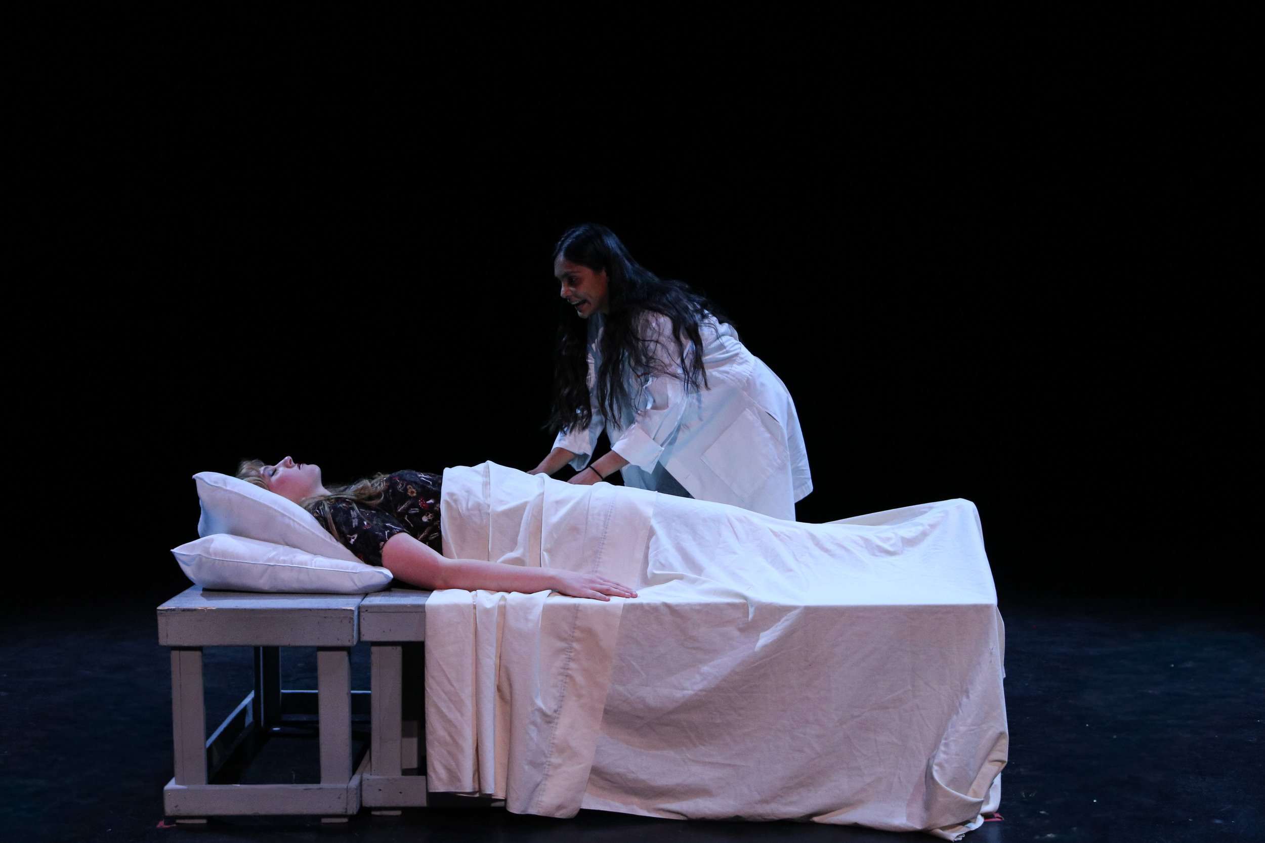 A woman lying in a hospital bed under white sheets, with another woman standing beside her in a dimly lit room with a blue glow.