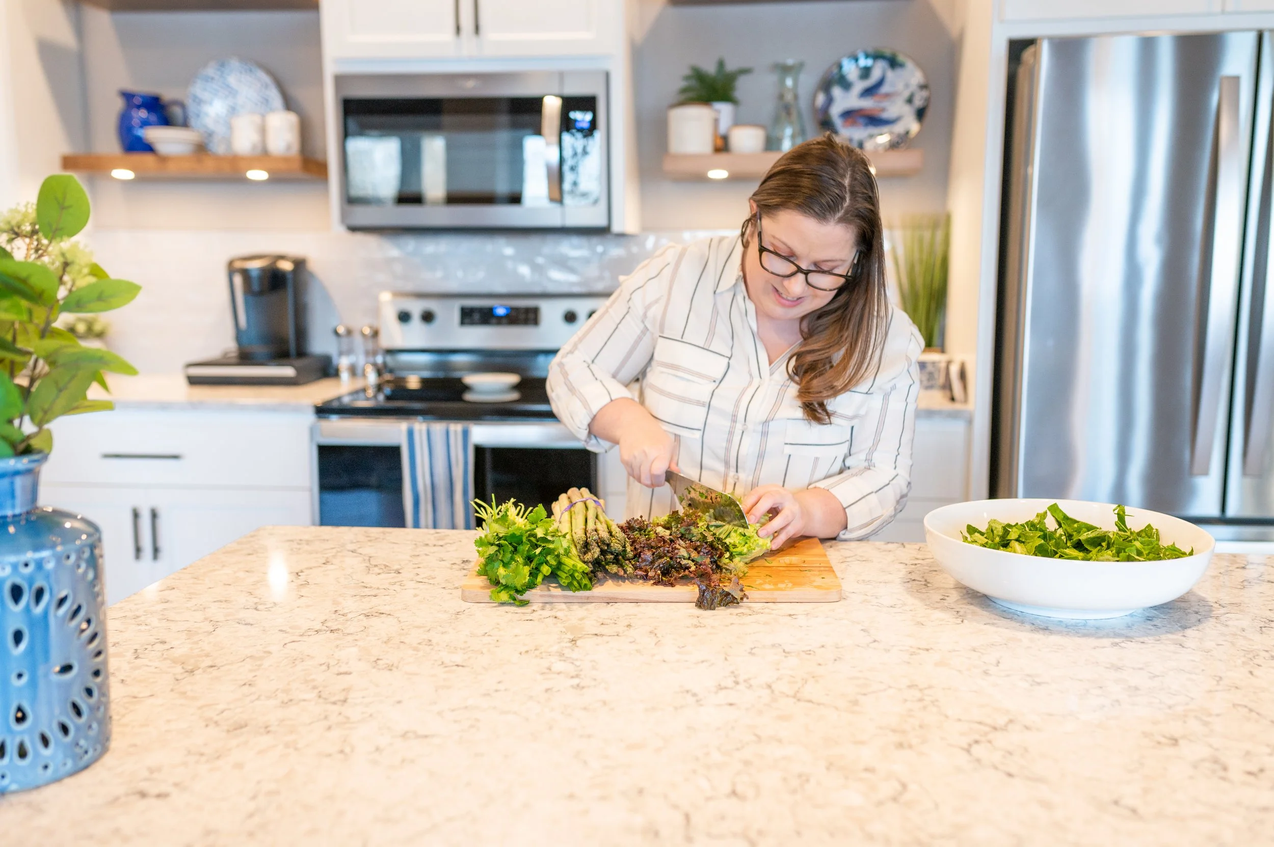 A woman with long brown hair and glasses preparing food in a kitchen, chopping leafy greens on a wooden cutting board. A large white bowl filled with greens is on the marble kitchen counter, with a blue decorative vase in the foreground. The kitchen has white cabinets, a microwave, a stainless steel refrigerator, and open shelves with decorative items.