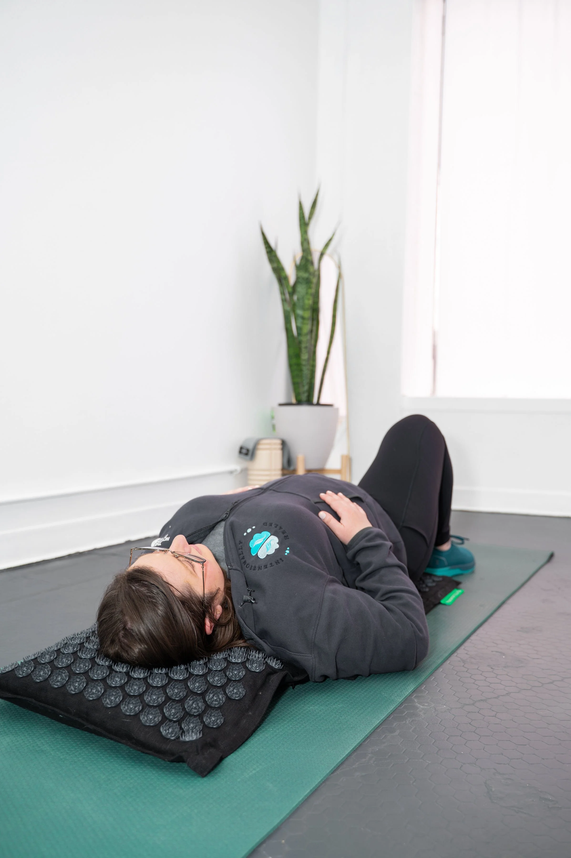 A person lying on a green yoga mat in a room, performing a guided relaxation or meditation exercise with a rolled black massage or acupressure mat beneath their upper back and head. The individual is wearing glasses and casual sportswear, with a hand resting on their abdomen. There is a white wall with a tall green plant, a window, and a small table with a phone and some books in the background.