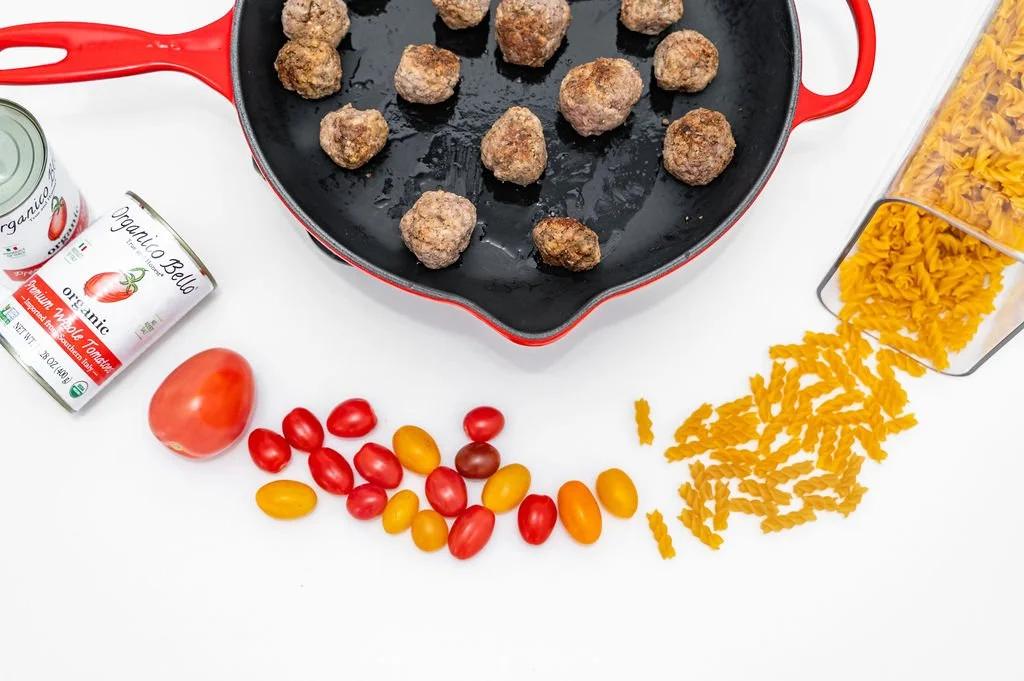 Uncooked meatballs in a red skillet, cherry tomatoes, a can of organic tomato juice, and cooked rotini pasta on a white surface.