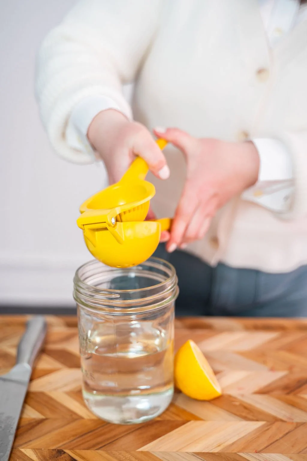 A person squeezing lemon juice into a glass of water using a yellow lemon squeezer, with a lemon wedge on a wooden surface and a knife nearby.