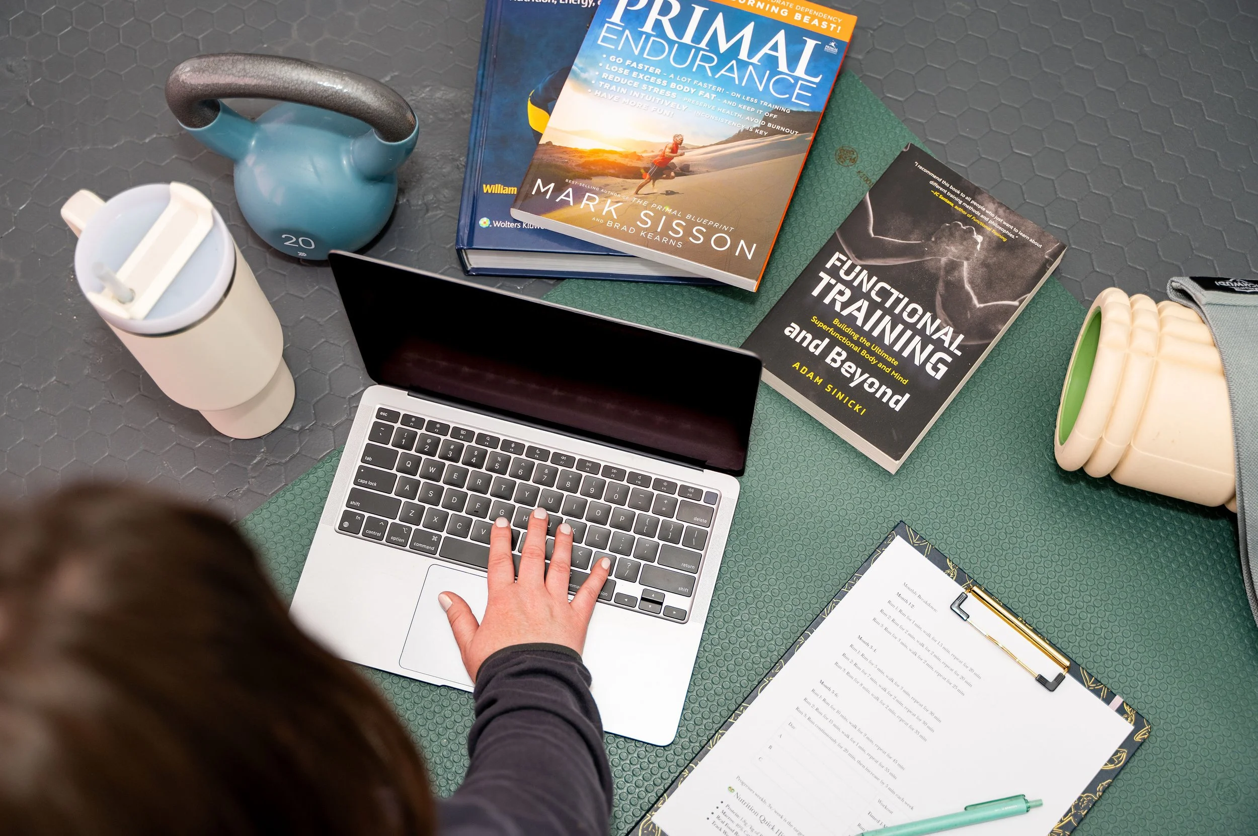 A person using a laptop with a hand on the keyboard, surrounded by books on fitness, work, and health, a kettlebell, a water bottle, a clipboard with papers, and a gray gym bag on a gray textured floor.