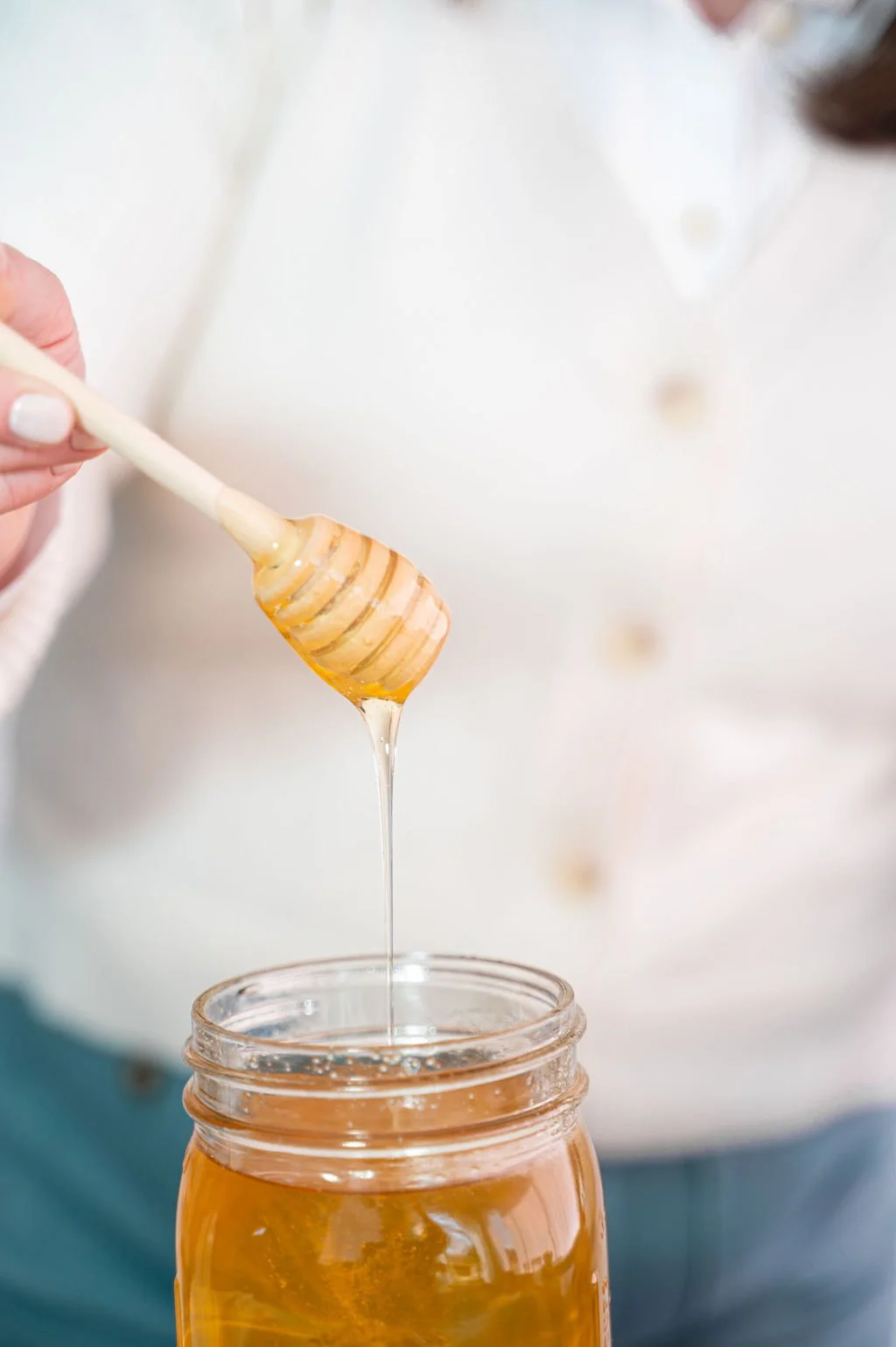 Close-up of a person holding a honey dipper dripping honey into a glass jar.