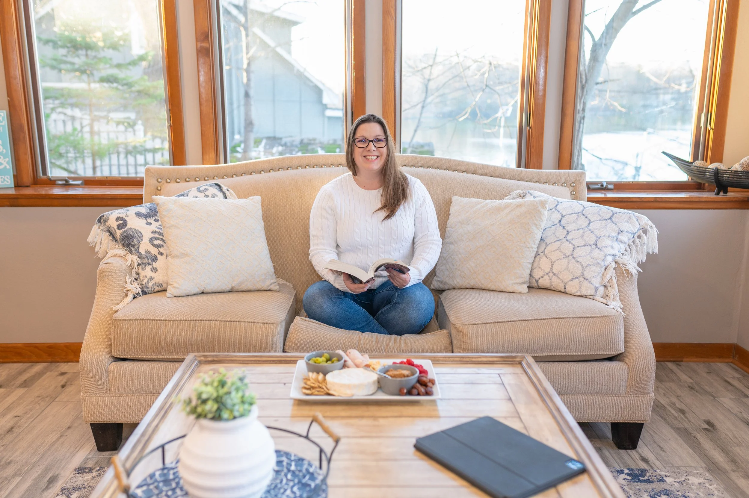 A woman with long brown hair, glasses, and a white sweater, sitting on a beige sofa with decorative pillows, smiling and holding a book. Behind her are large windows showing a snowy outdoor scene with trees. A wooden coffee table in front of her holds snacks and drinks, with a decorative vase of greenery and a black leaf-shaped plate nearby.