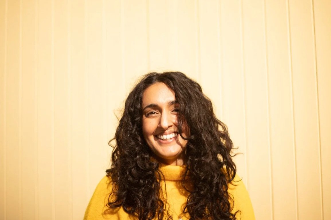 Manpreet Dhesi a smiling woman with long curly hair wearing a yellow sweater standing in front of a yellow panelled wall.