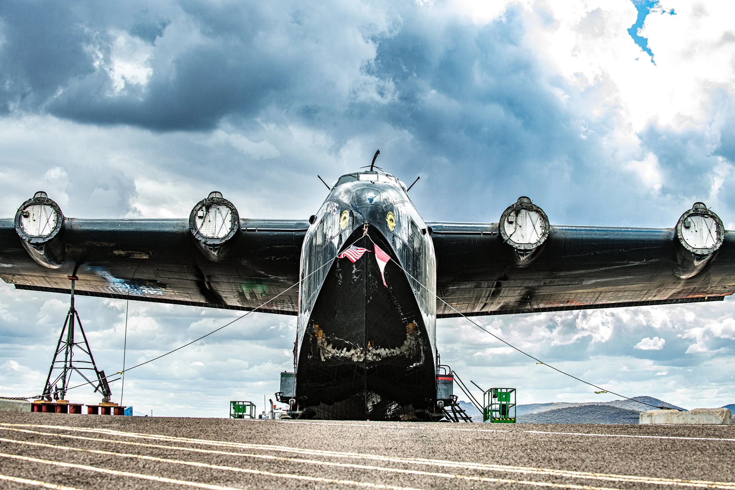 A large black military aircraft is parked on the tarmac under a cloudy sky. It has four engines on each wing and flags hanging from the front of the plane.