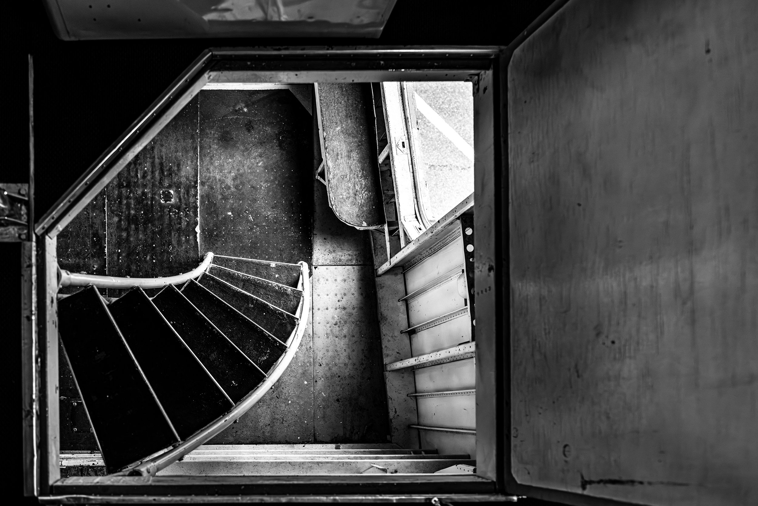 Black and white aerial view of a spiral staircase inside a building, viewed from above, with walls and floor visible.