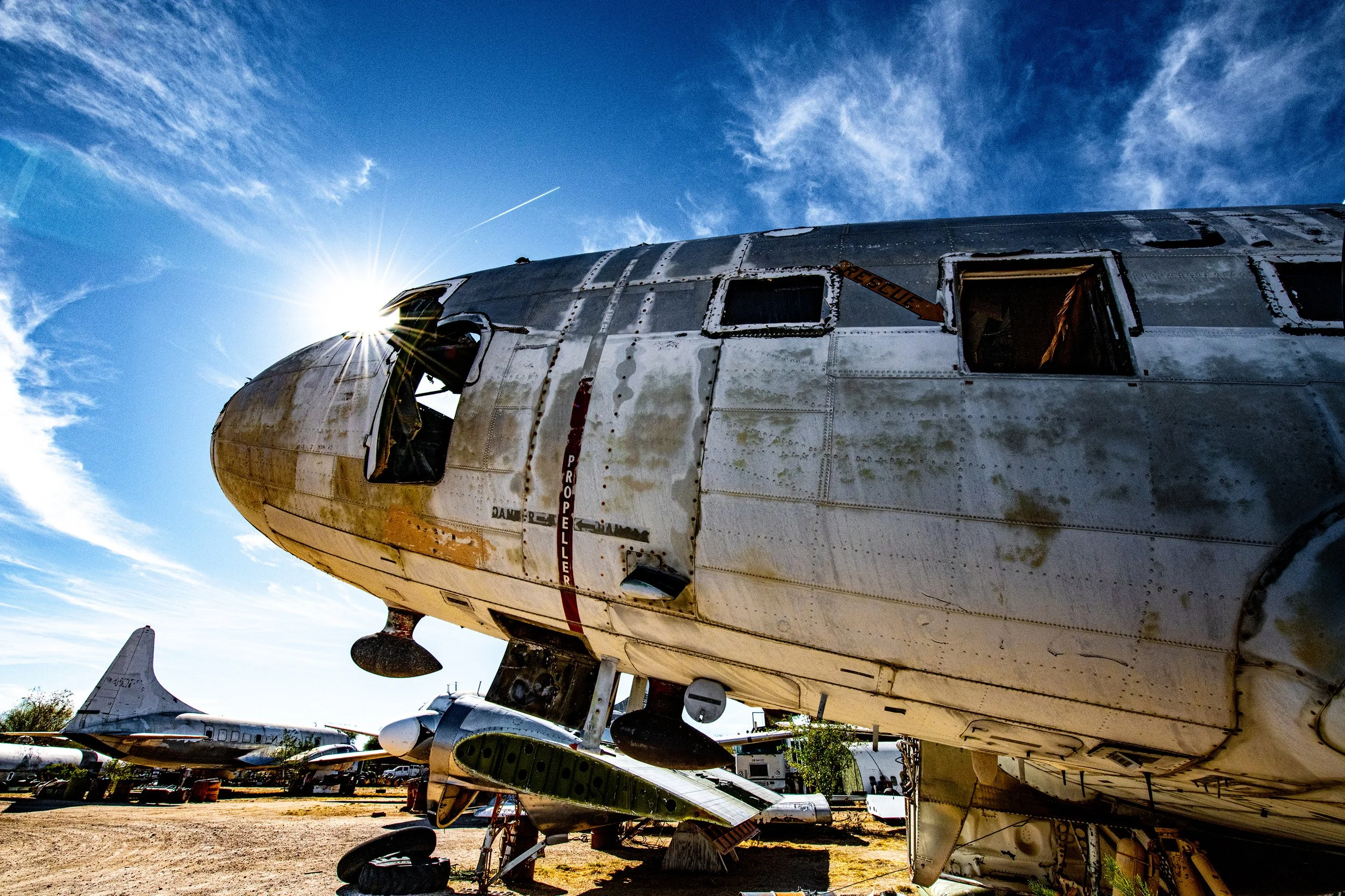 Upside-down, abandoned military aircraft in a scrapyard under a blue sky with clouds. The sun shines behind the aircraft, creating lens flare.
