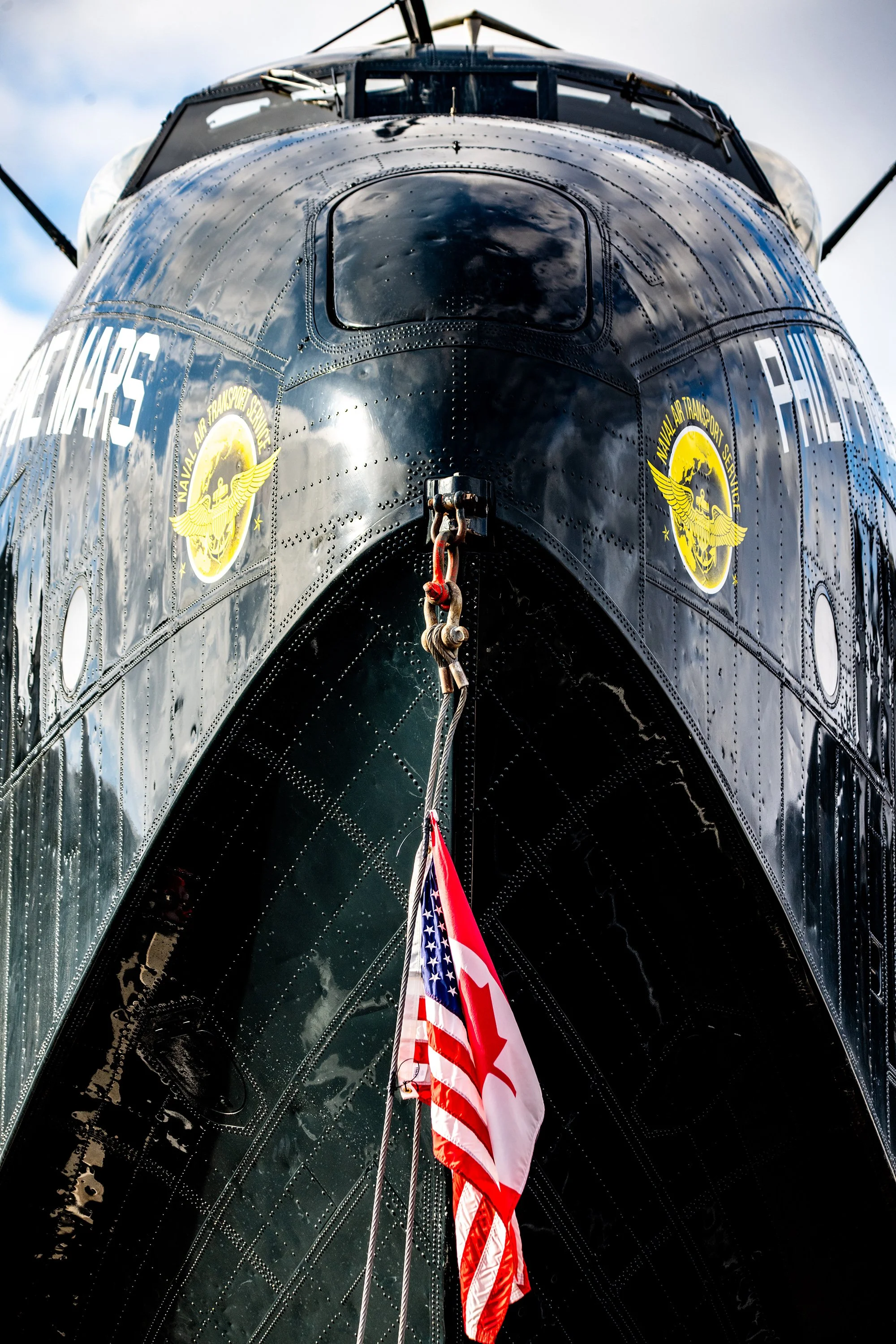 Close-up view of the bow of a navy submarine with a Canadian and American flag hanging in front, Navy Allied Transport Service insignia on the sides, and a reflective black hull.