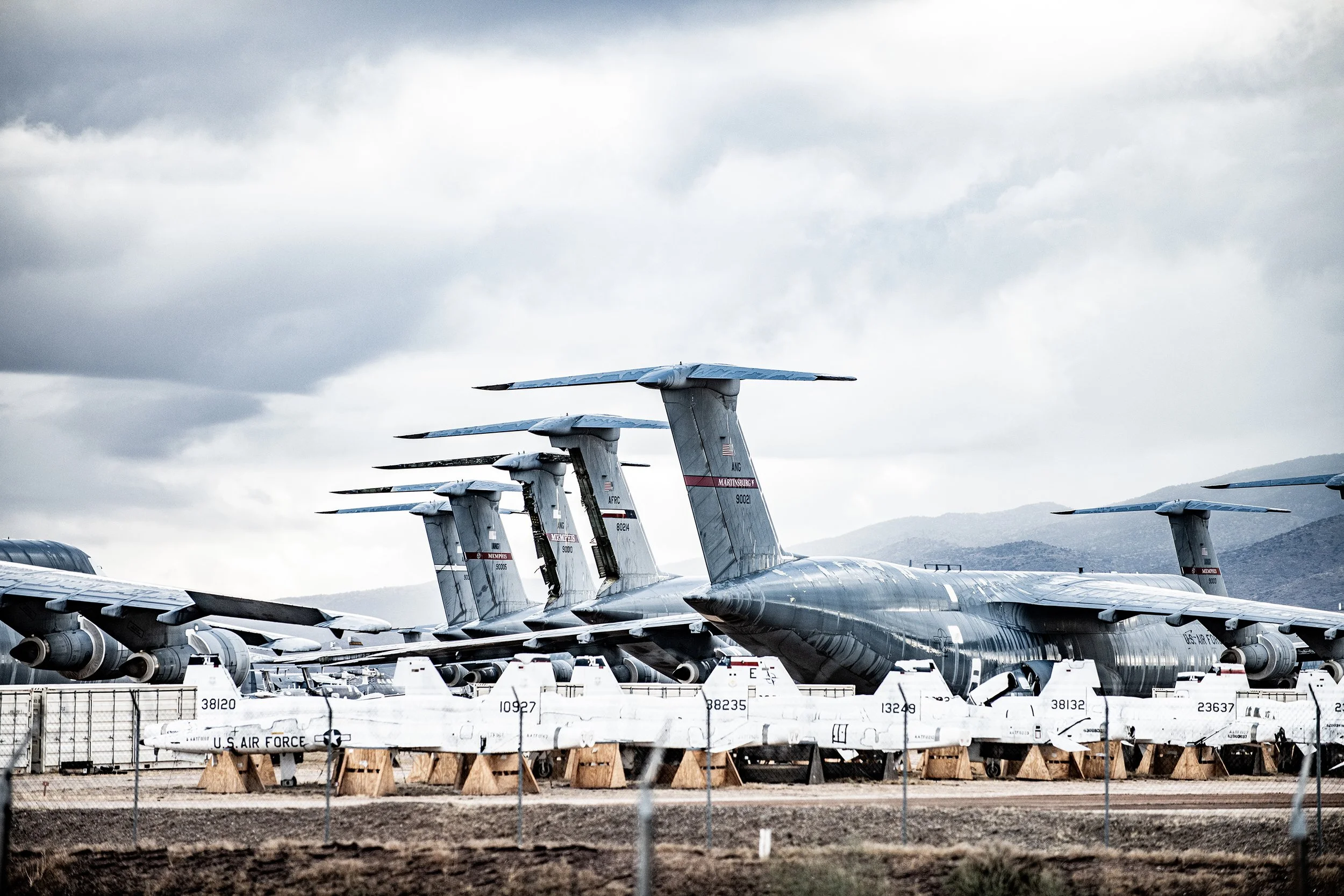 Multiple U.S. Air Force military aircraft parked on a tarmac under cloudy sky, with mountains in the background.
