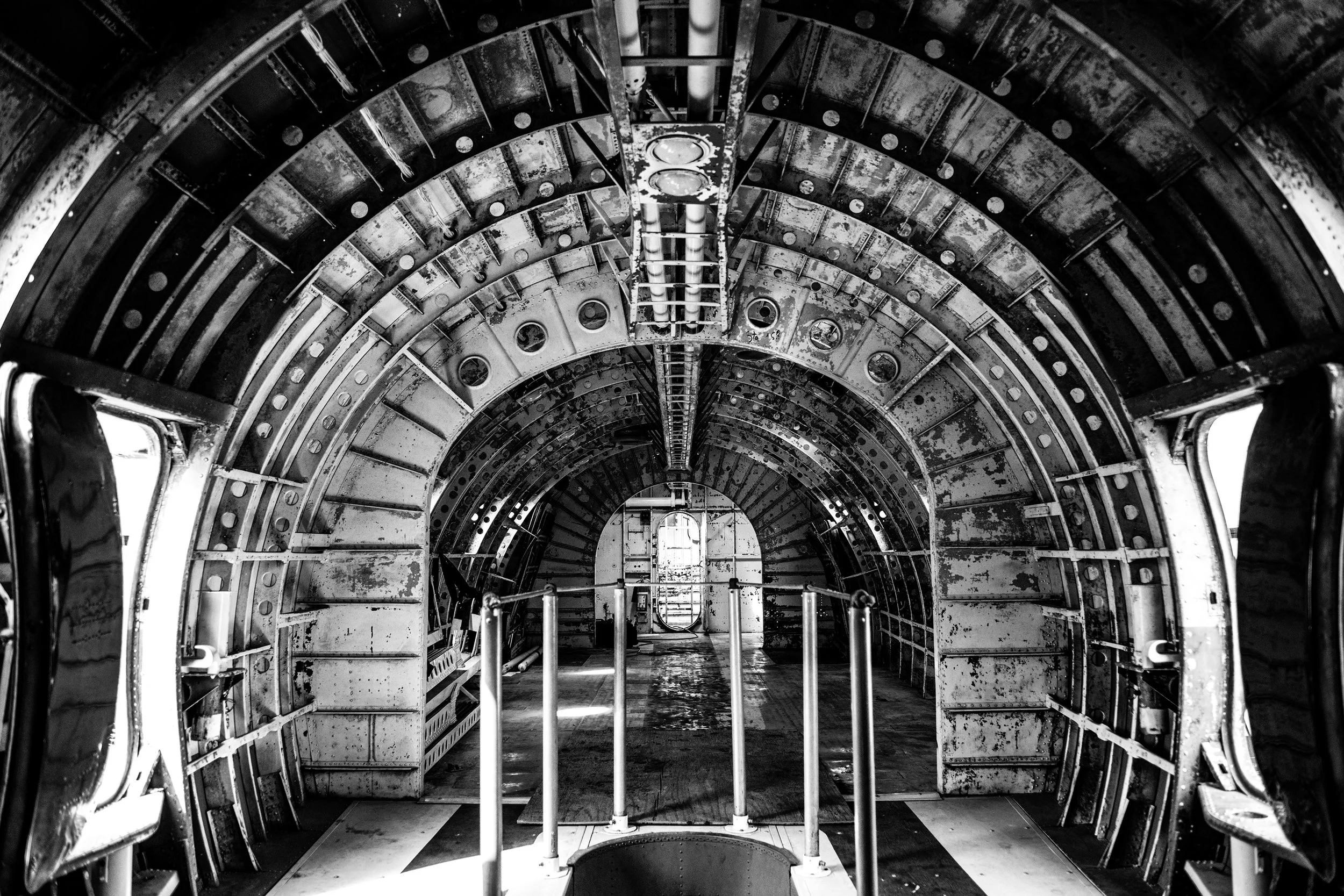 Black and white photo of the interior of a large, cylindrical metallic structure, possibly inside a submarine or spacecraft, with curved walls, pipes, and support beams, leading to a door at the far end.