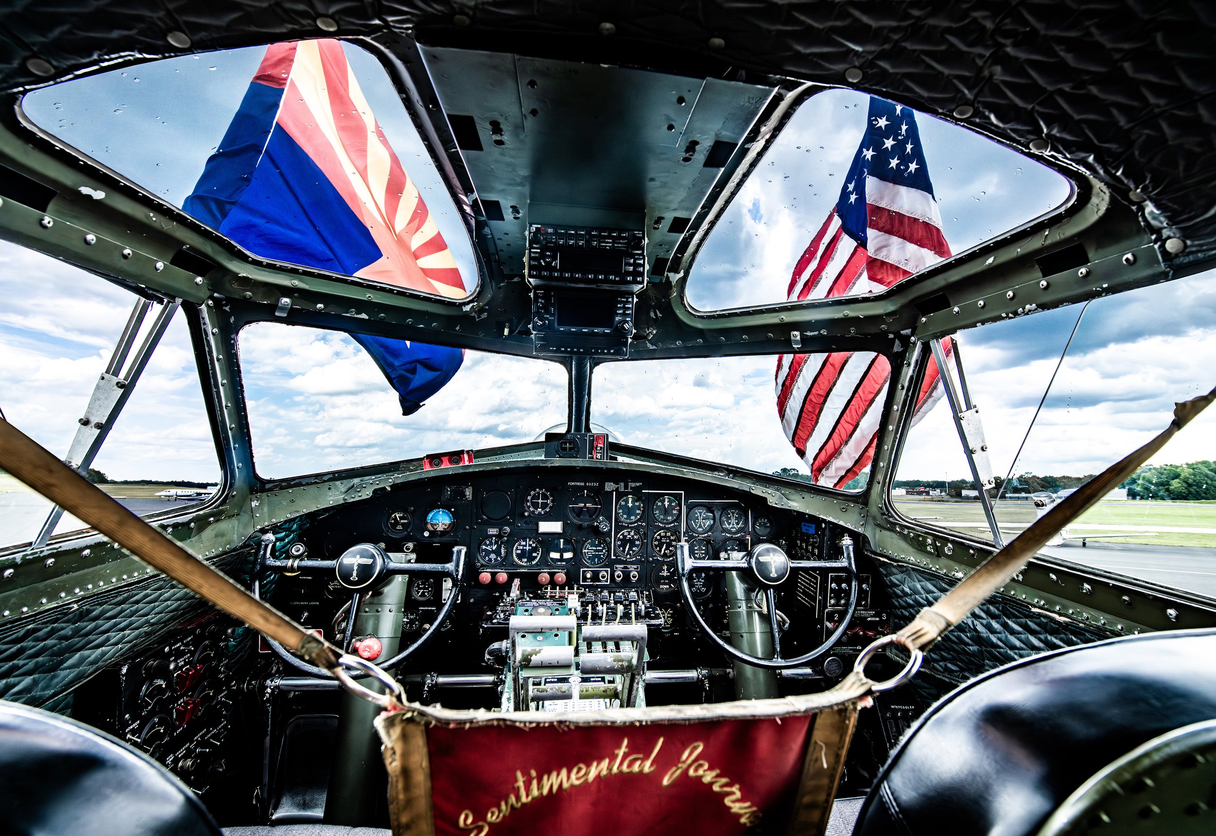 Interior of a vintage airplane cockpit with multiple gauges and controls, displaying two American flags and one other flag through the windows.