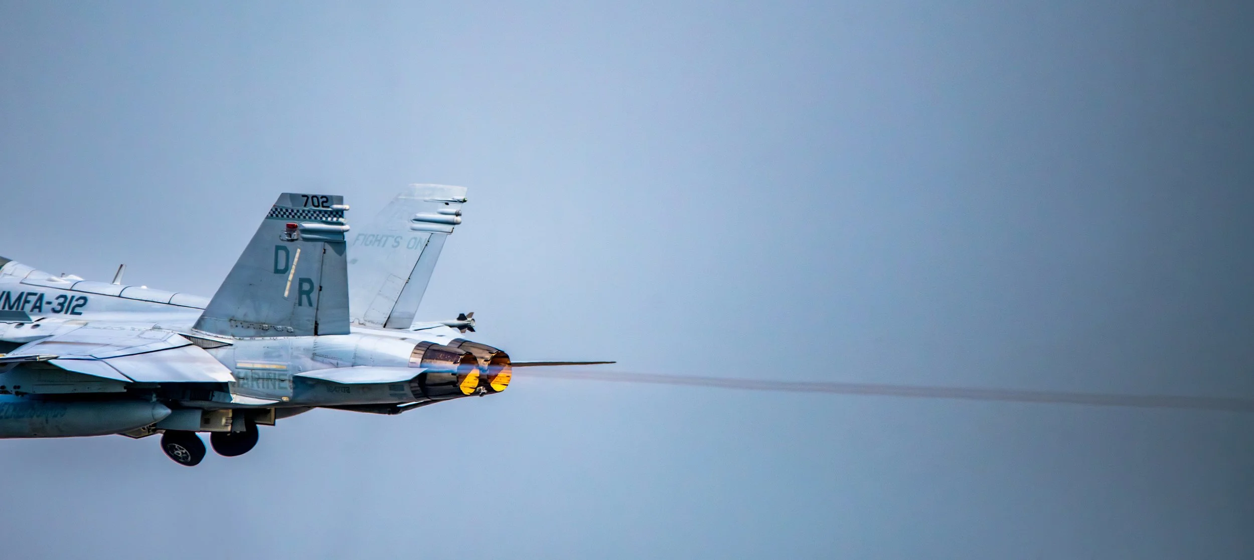 Military jet aircraft flying at high speed with afterburners activated, leaving a trail of jet exhaust against a cloudy sky.