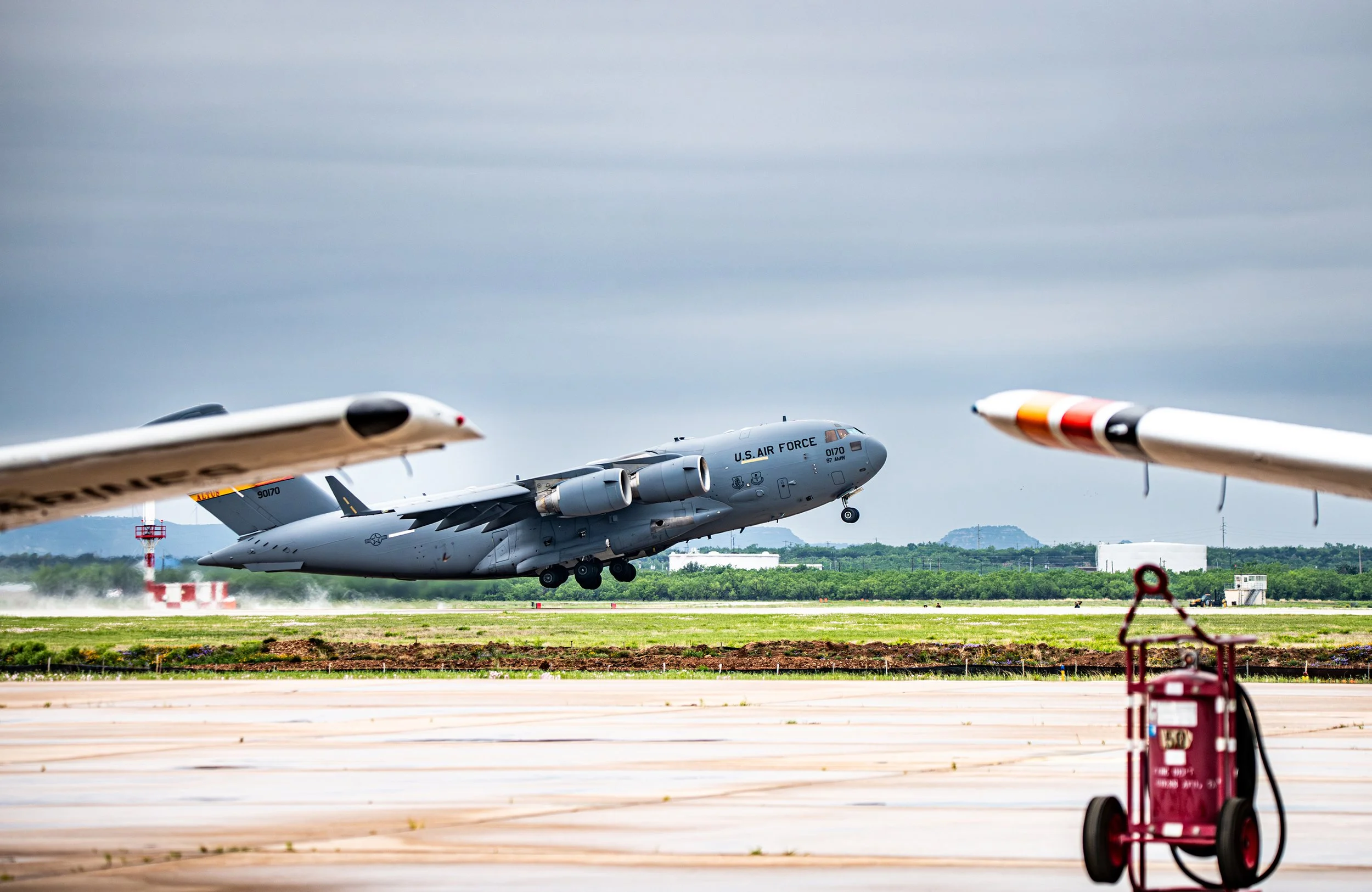 A U.S. Air Force transport plane taking off from an airport runway during daytime. The aircraft is gray and has four engines, with two winger and two engine nacelles on the fuselage. The foreground shows parts of two other aircraft wings and a red fi