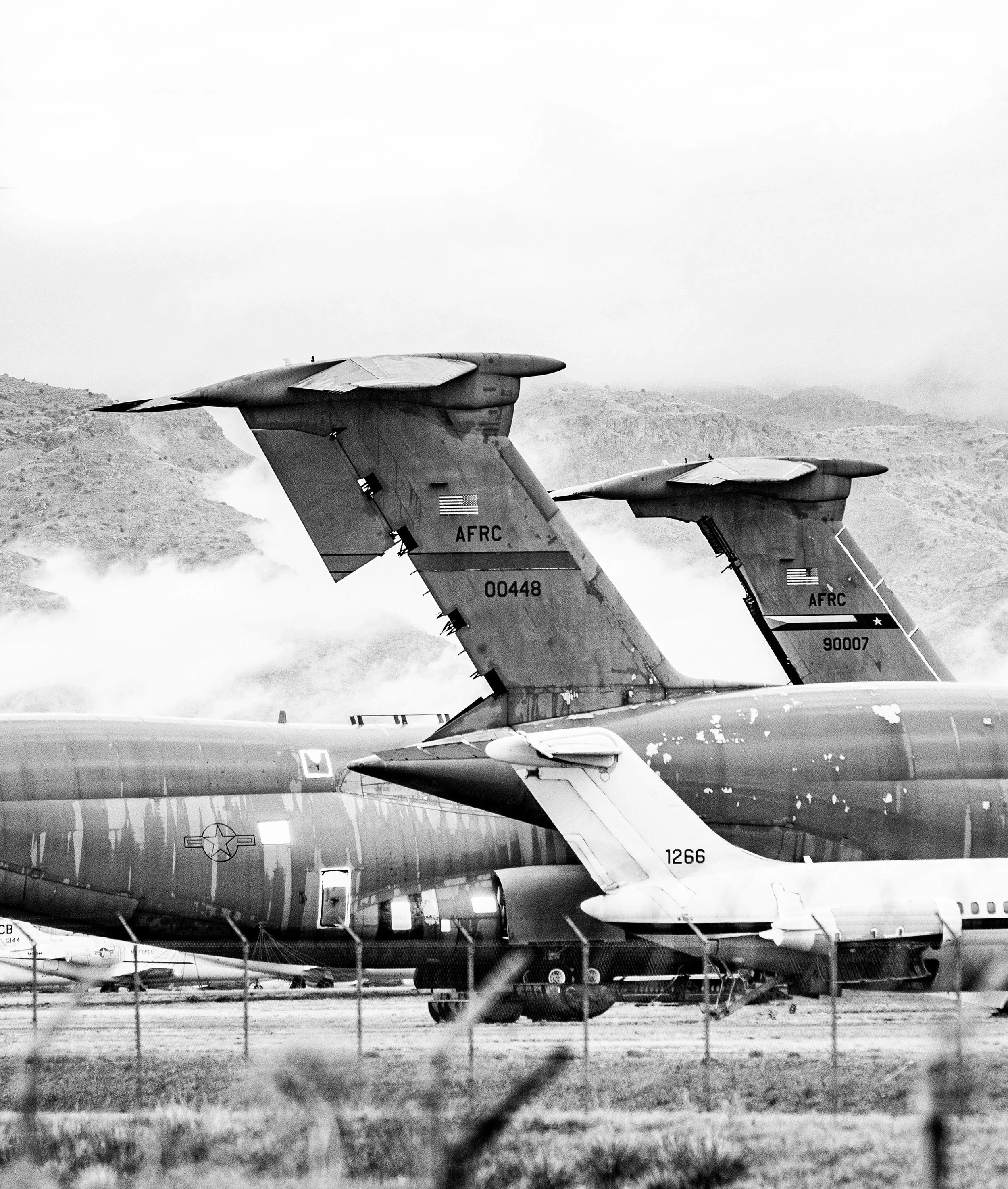 Two military fighter jets parked on the ground near a chain-link fence with mountains in the background.