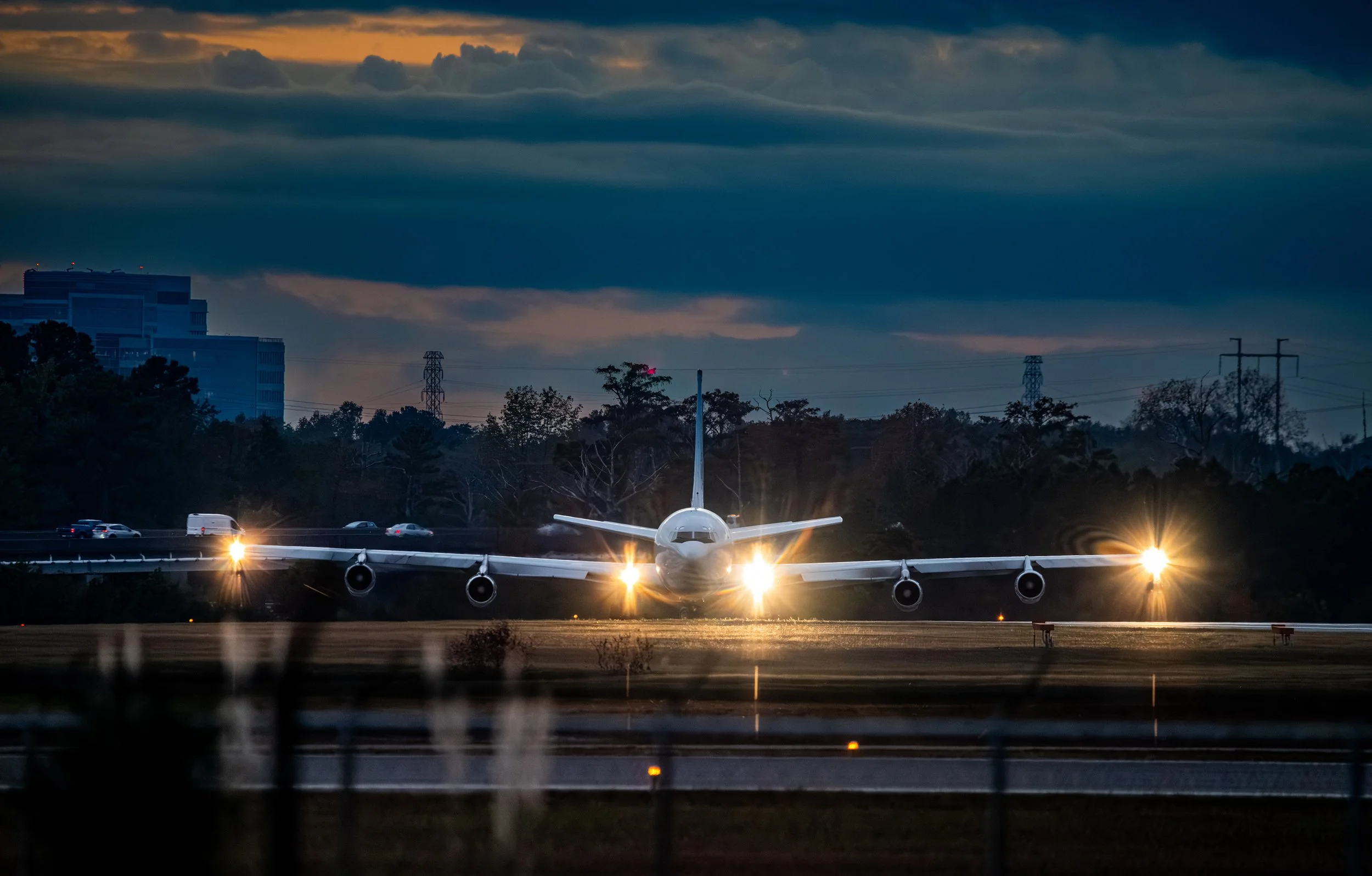 An airplane on a runway with its lights on during dusk, with a cloudy sky and trees in the background.