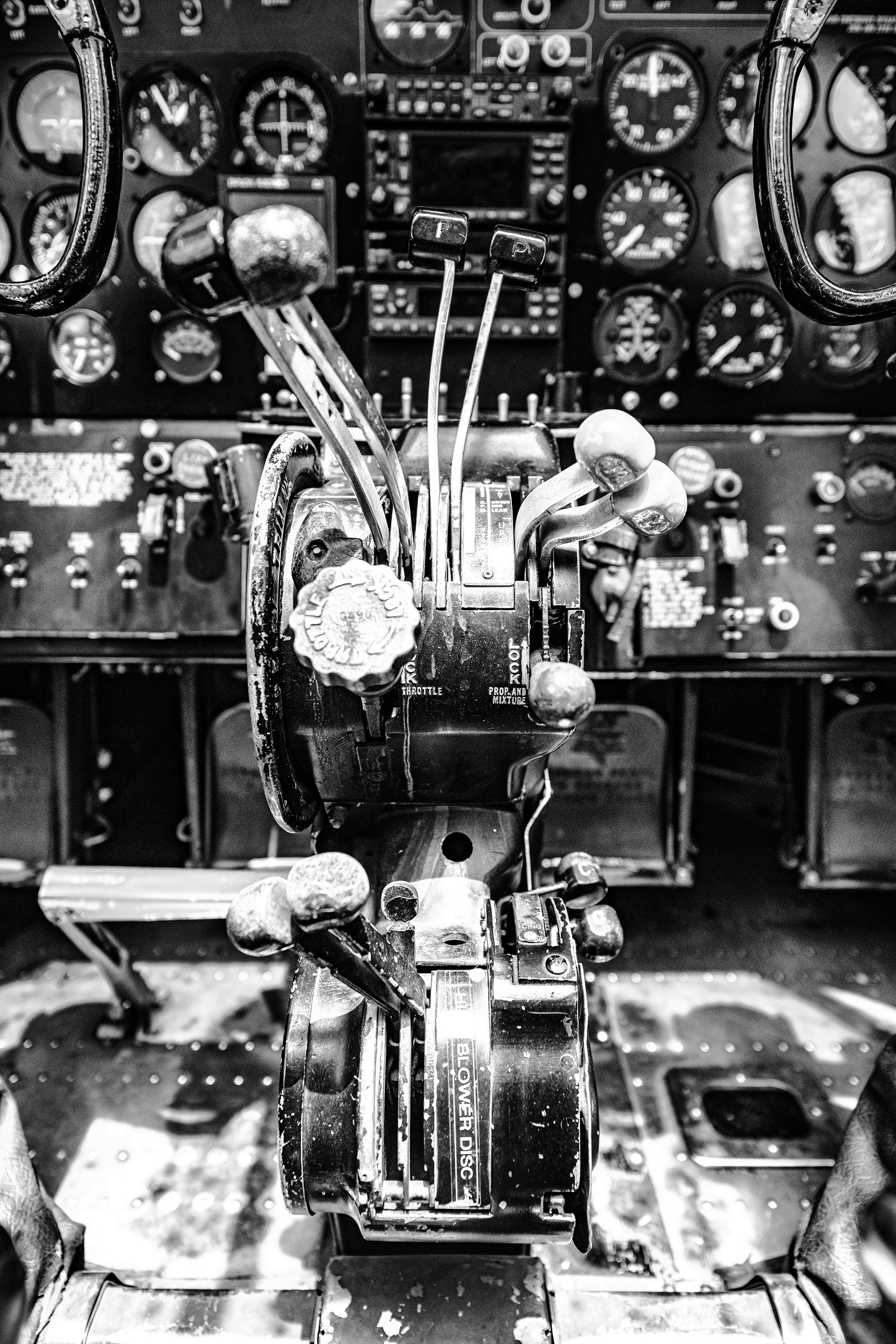 Black and white photo of the cockpit controls of a vintage airplane, featuring a central control stick with various levers and knobs, surrounded by numerous gauges and instruments.