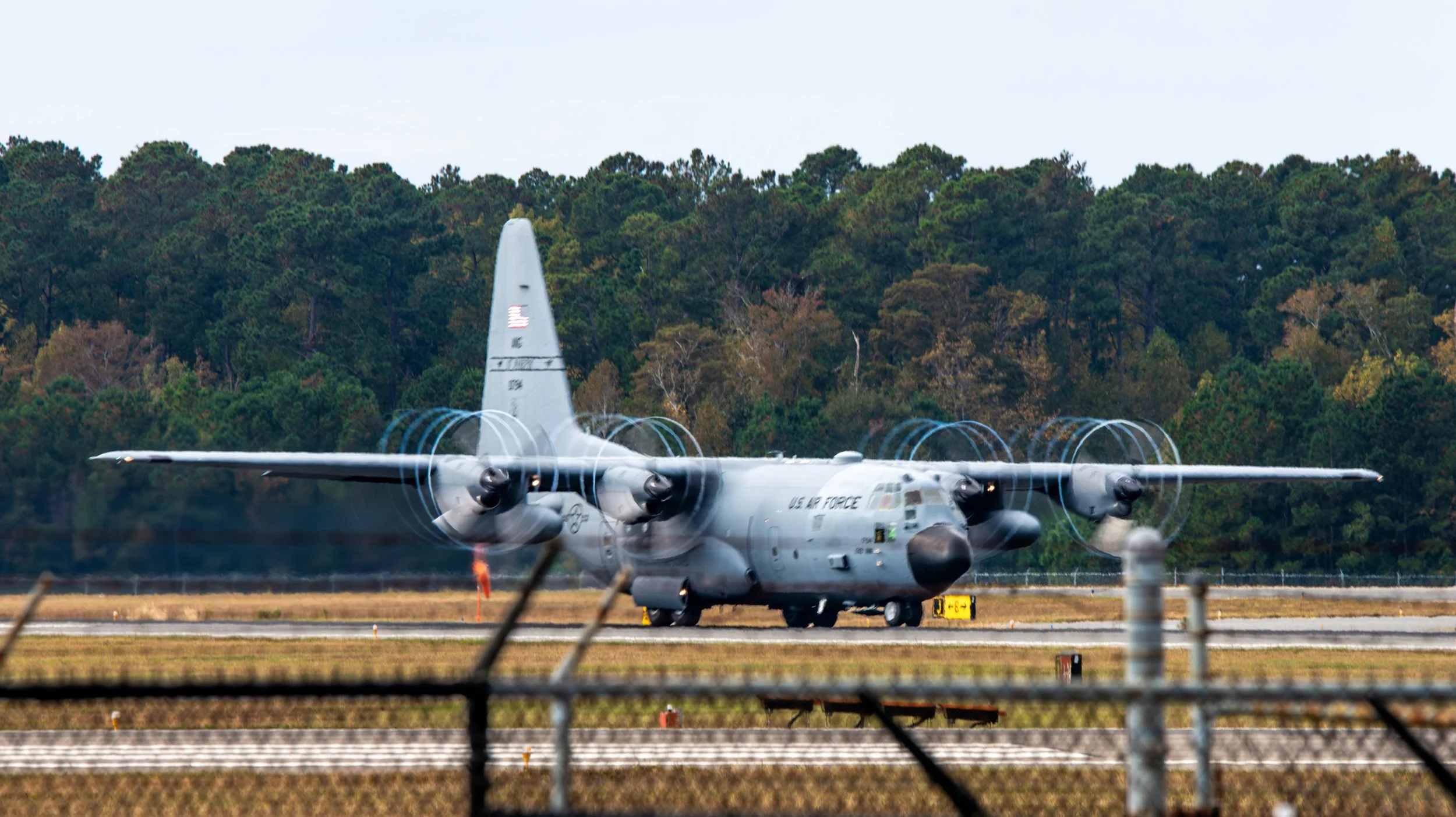 A U.S. Air Force military airplane on the runway with four propellers, surrounded by a chain-link fence and trees in the background.