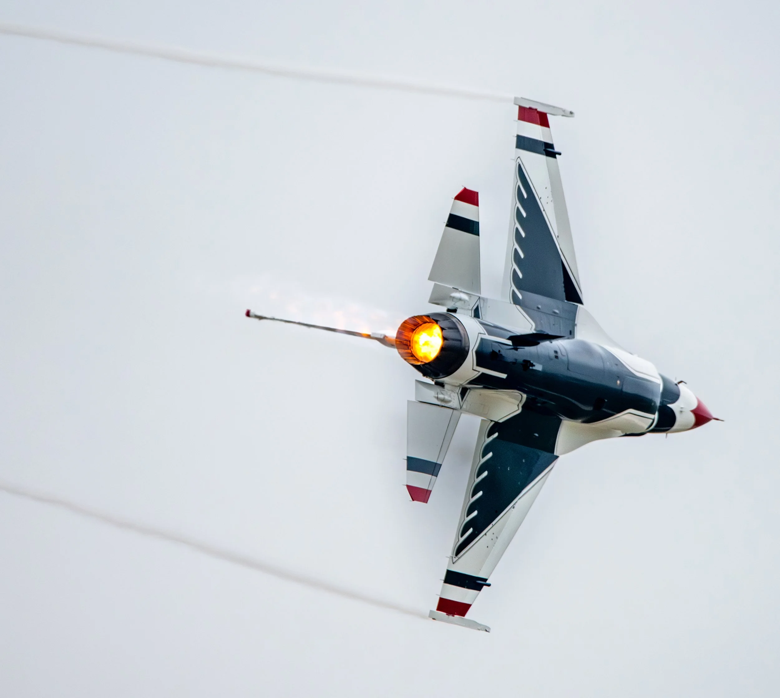 A fighter jet plane in mid-flight with afterburner engaged, emitting a bright flame, against a cloudy sky.