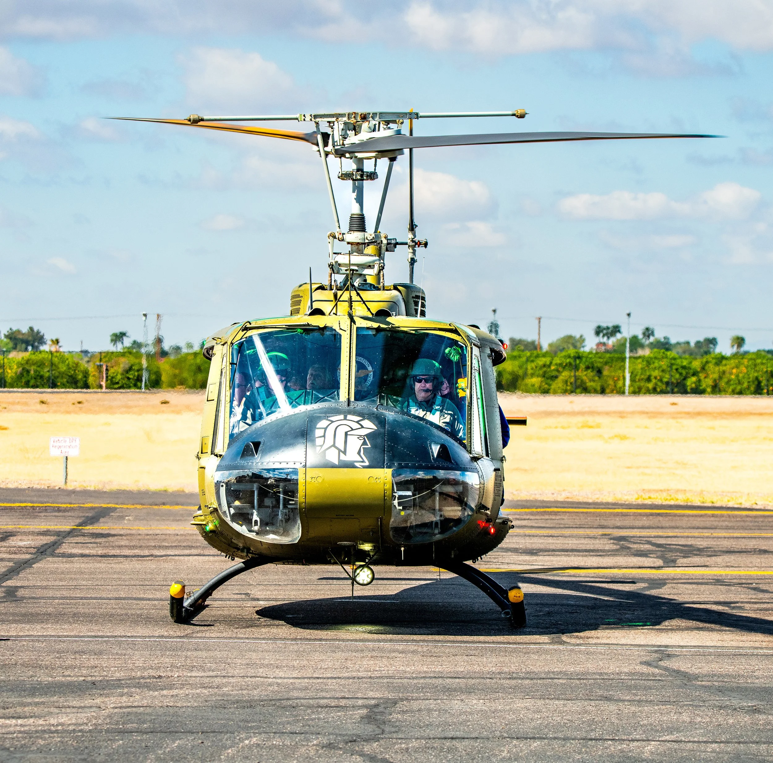 A military helicopter with a black and yellow color scheme parked on an airfield, with pilots visible inside the cockpit against a background of blue sky and open field.