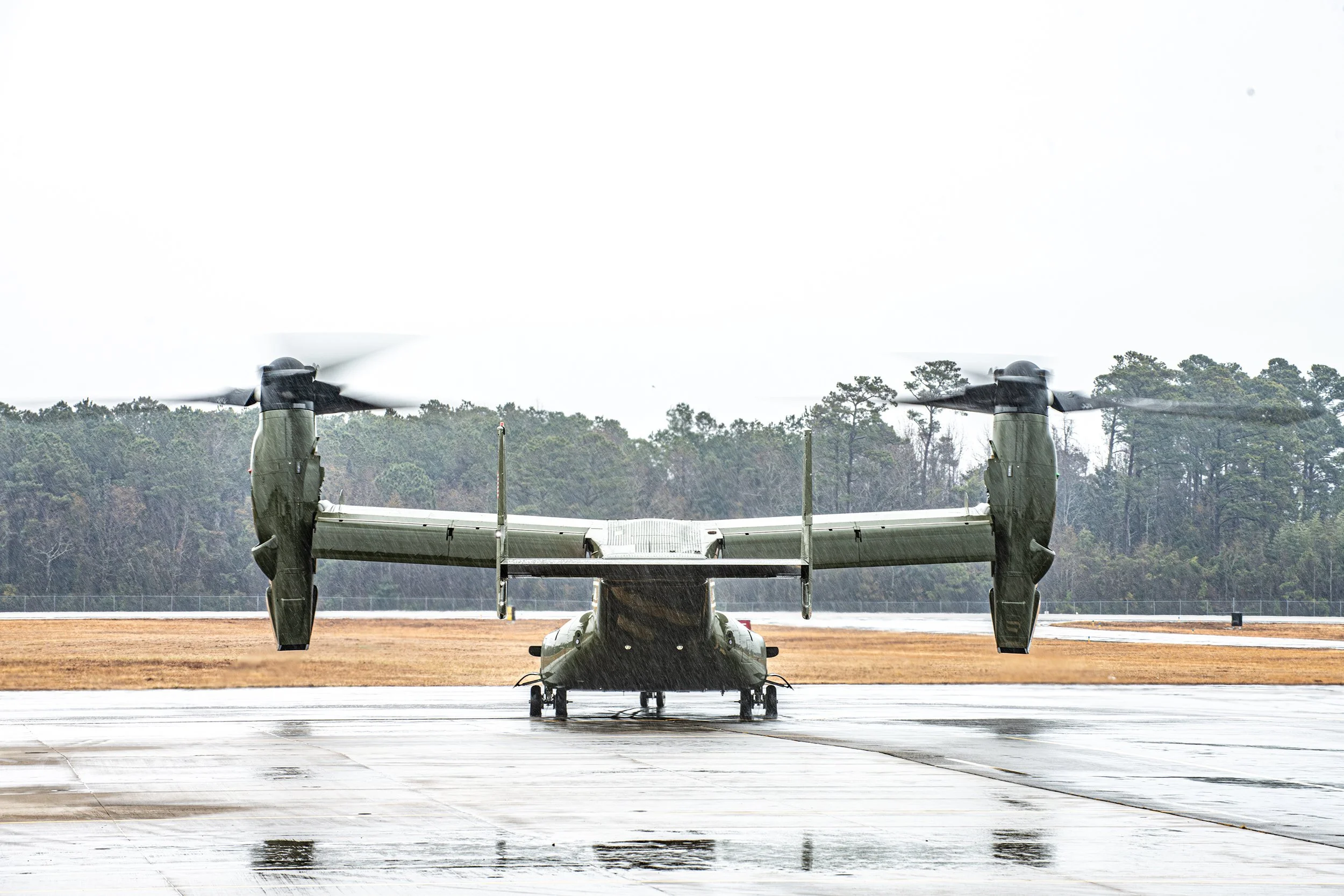 A military aircraft with vertical takeoff and landing capabilities on wet tarmac, with rain and a cloudy sky, and forested area in the background.