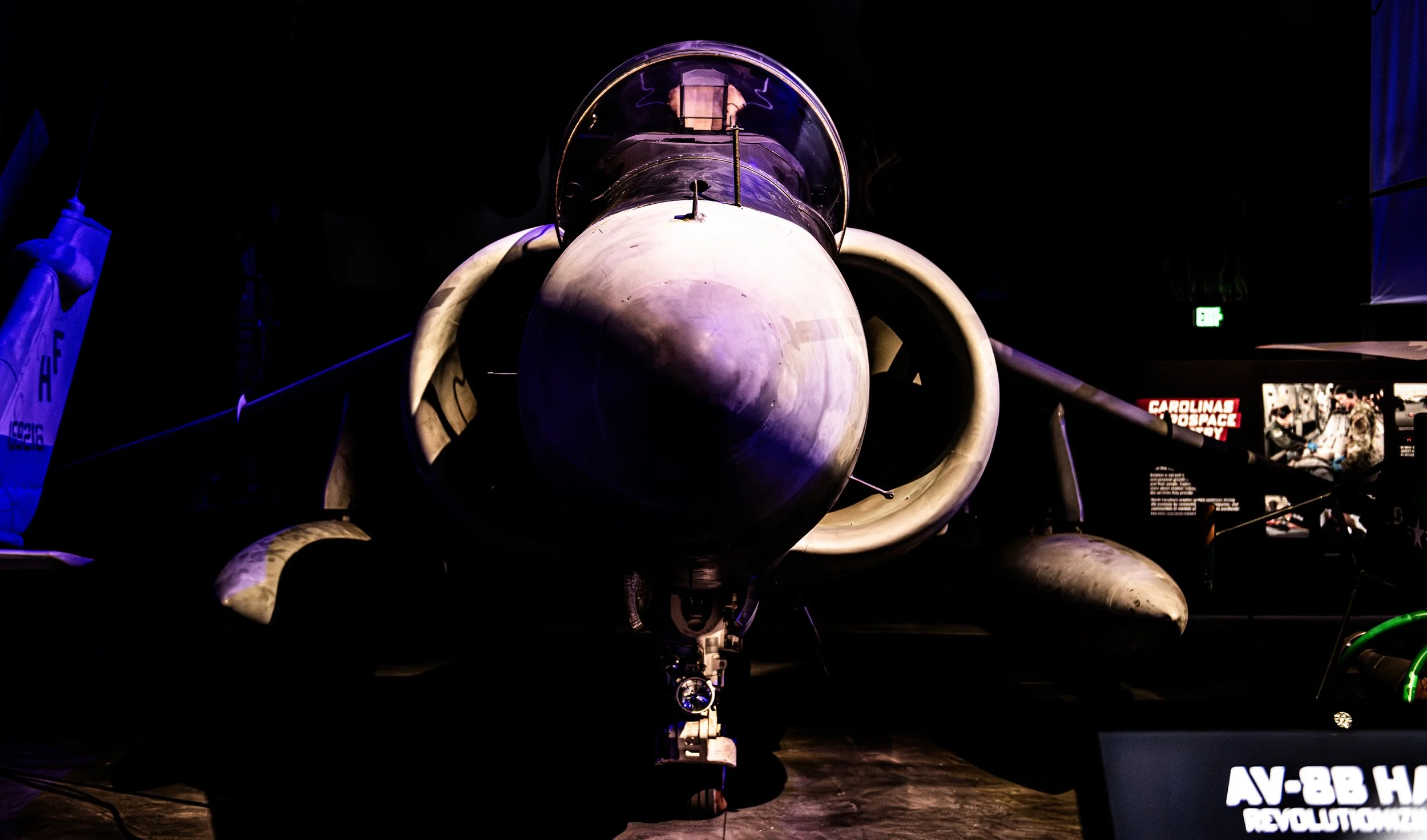 Front view of a military fighter jet on display in a dimly lit museum or exhibit, with signage and informational panels visible in the background.