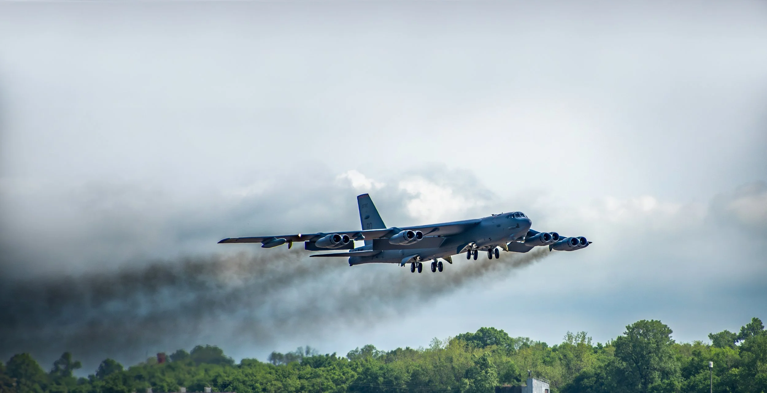 A large military aircraft flying low over a forest, emitting dark smoke from its engines.