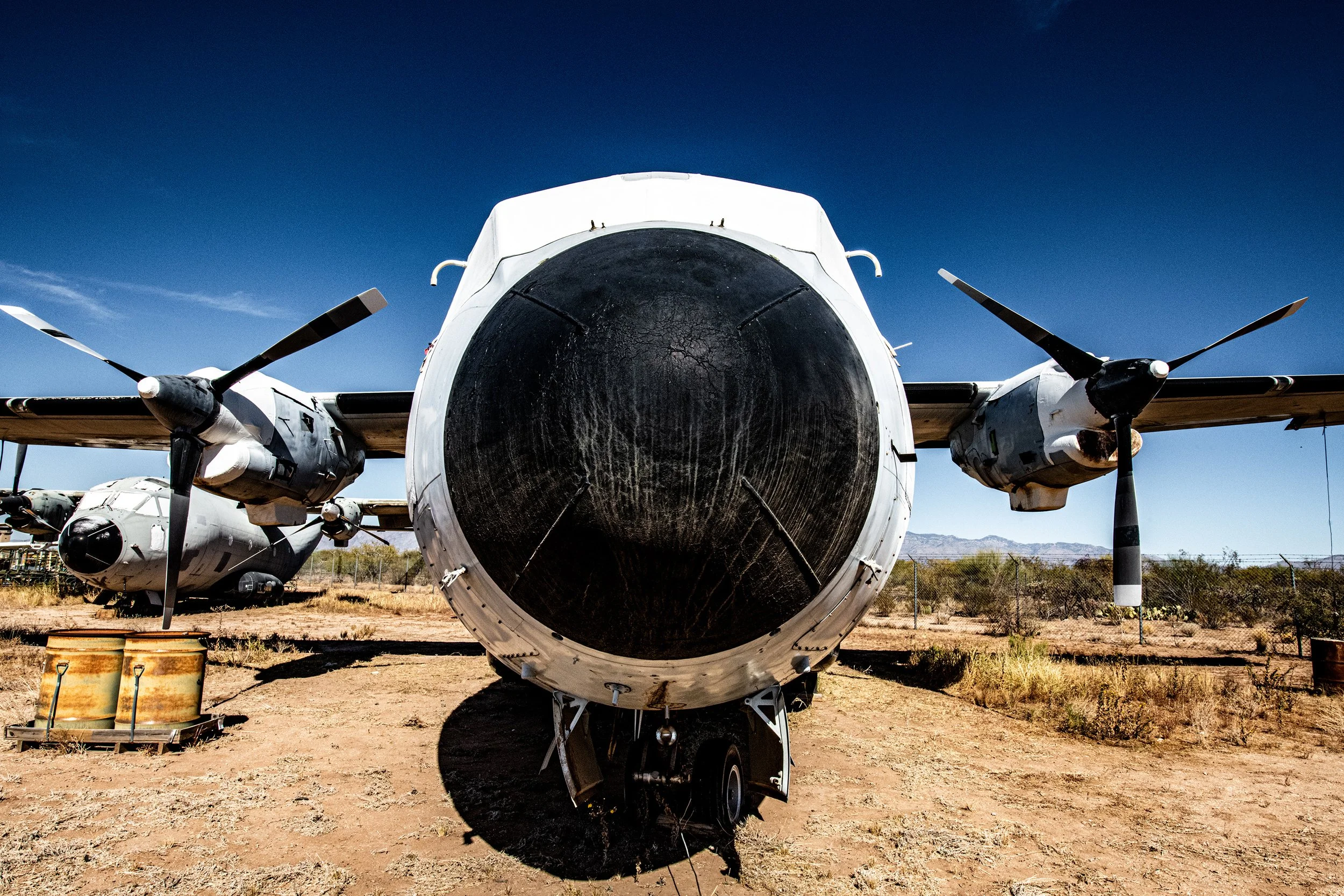 Front view of an aircraft with a large, blackened nose cone, parked on a dirt surface under a clear blue sky.