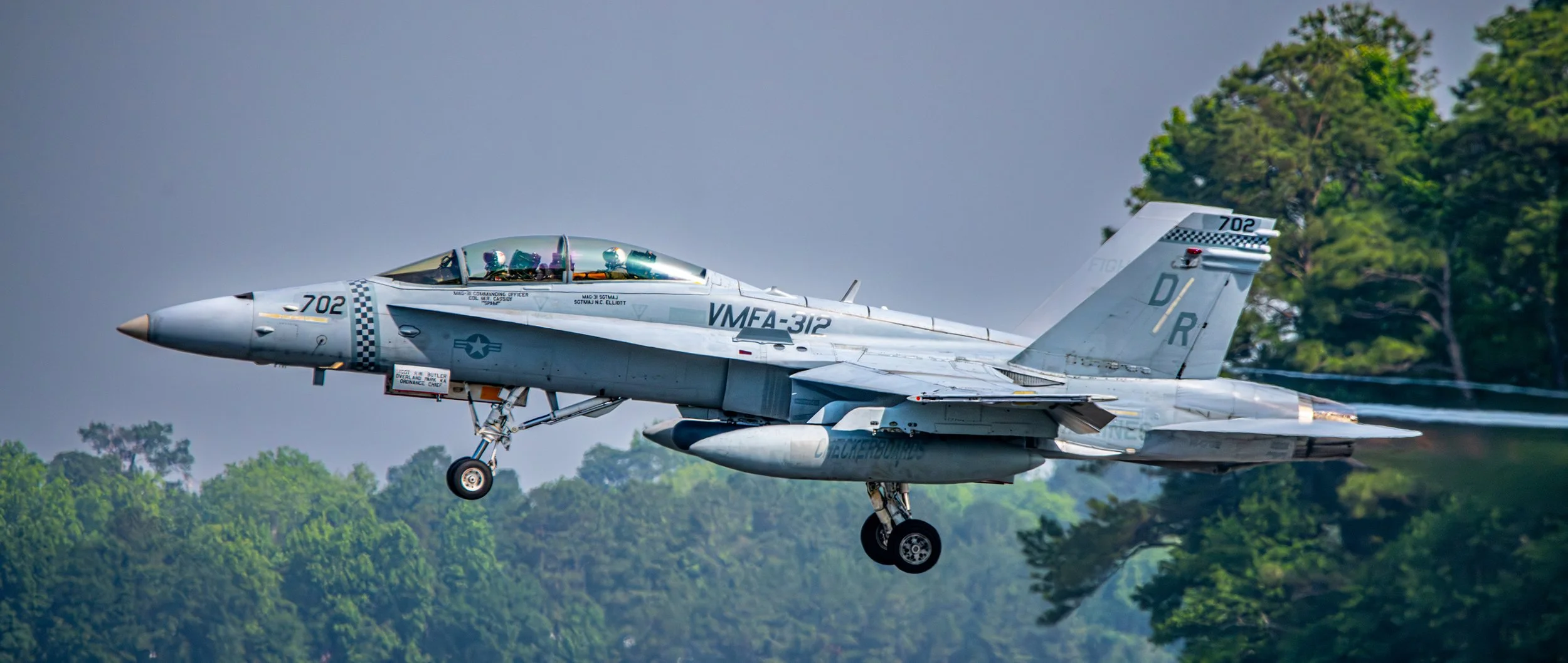 Military fighter jet airplane flying low over green trees with afterburners active.