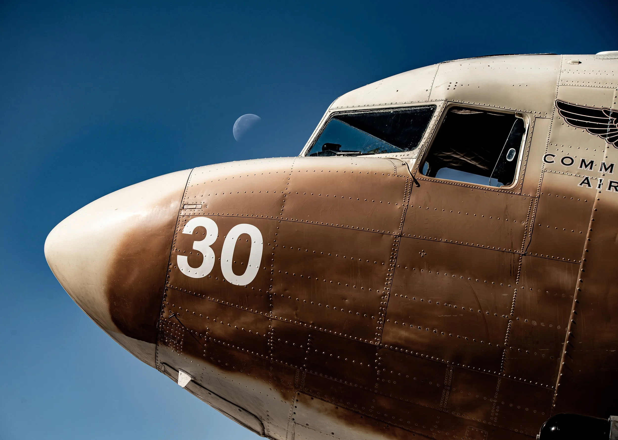 Close-up of a vintage military aircraft nose with the number 30, part of the cockpit, and an aircraft insignia on the side, against a clear blue sky featuring a visible crescent moon.
