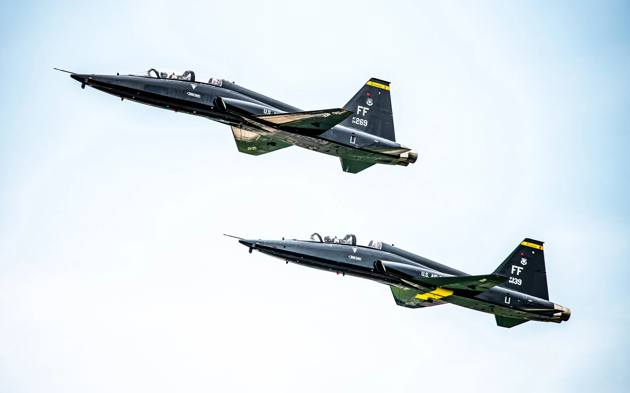Two black fighter jets flying in formation against a light blue sky.