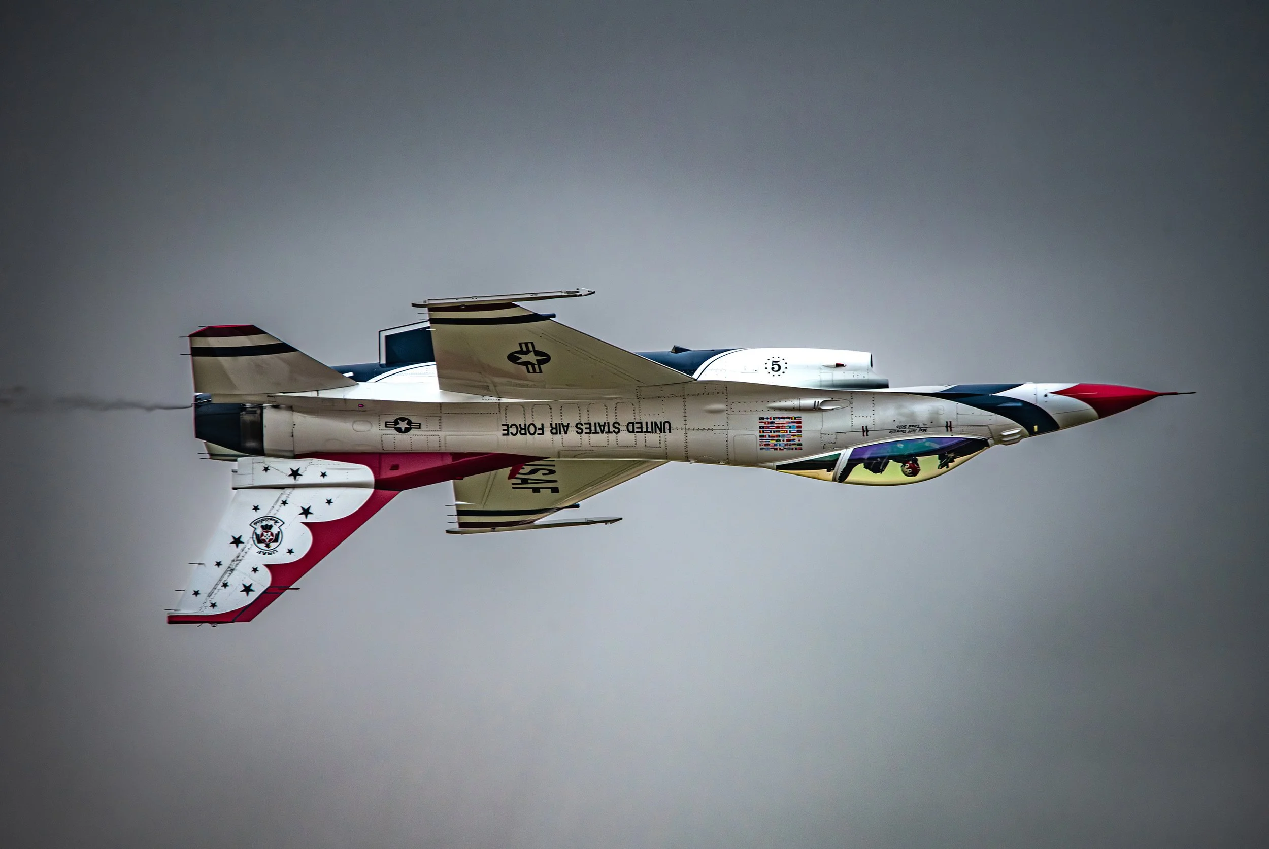 A fighter jet flying upside down against a gray sky, with patriotic red, white, and blue paint and markings indicating it is from the United States Air Force.