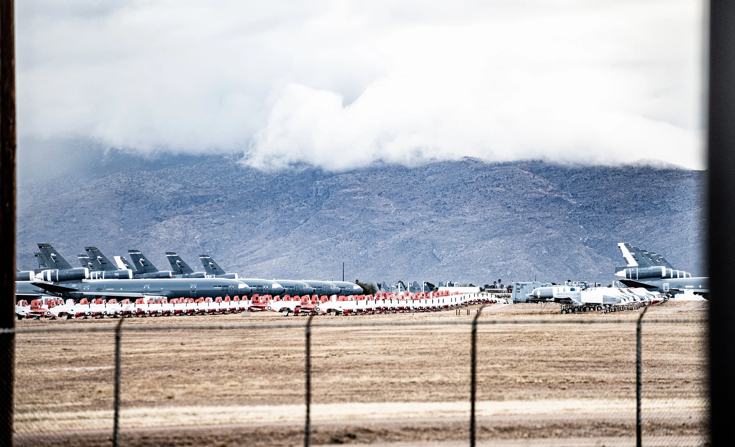 Military planes parked on an airfield with mountains and cloudy sky in the background, viewed through a fence.
