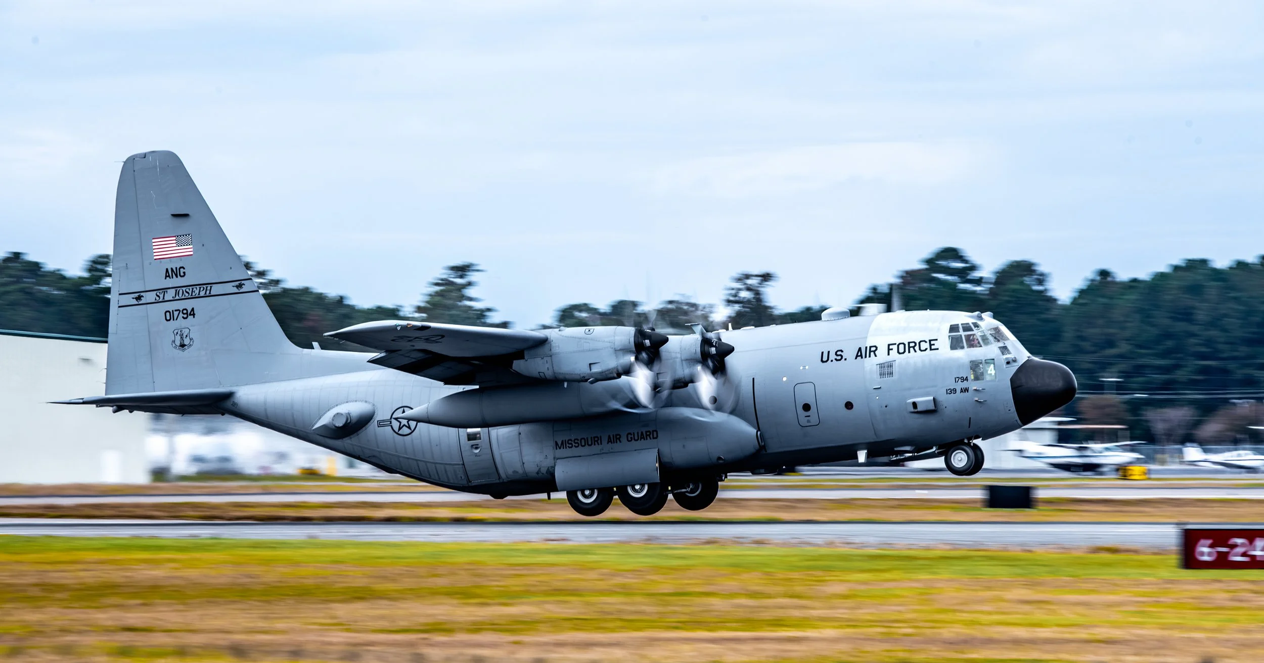 A gray U.S. Air Force Lockheed C-130 Hercules military aircraft taking off from a runway, with trees and airport facilities in the background.